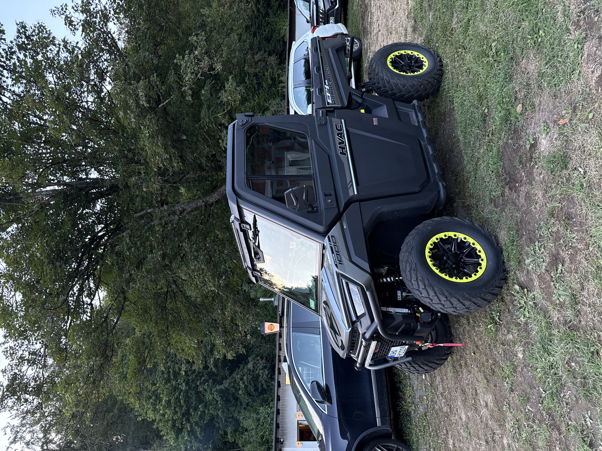 The image shows a dark grey UTV with yellow accents on its wheels parked on a patch of grass. The UTV has a closed cab with windows and a roof. In the background, there are trees and a building with a sign. To the left of the UTV, a black SUV is parked. The scene appears to be outdoors, possibly in a rural or semi-rural area. The UTV has a license plate that reads "OHA H1110". The building in the background has a sign that appears to be an advertisement for beer. This scene is located in the town of Walkenried, Germany.