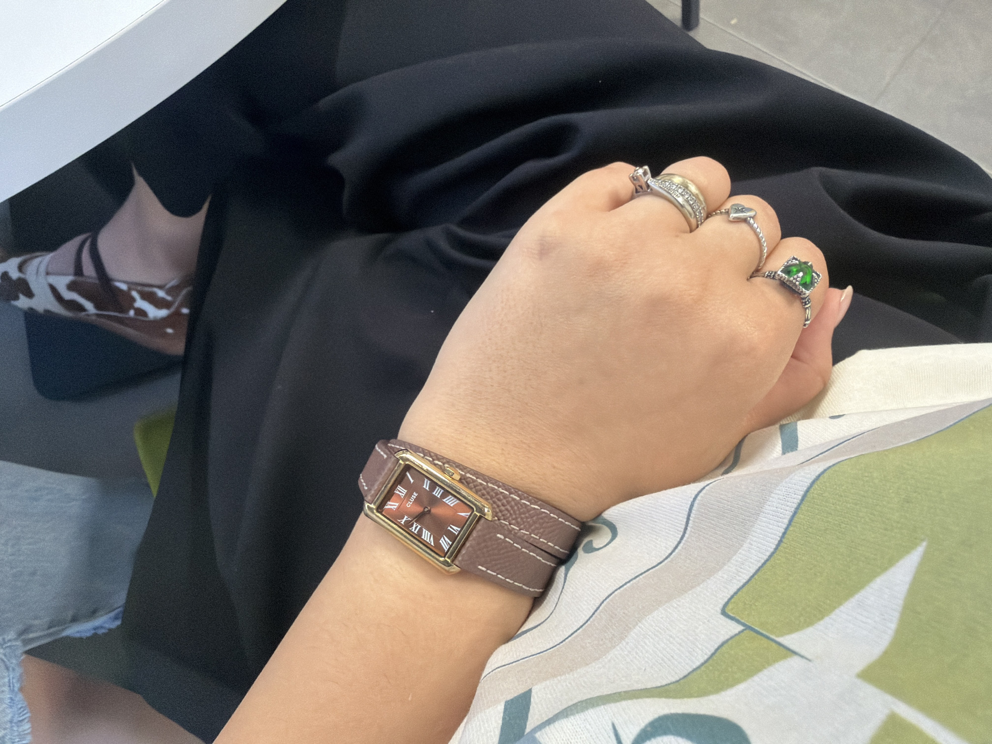 The image shows a person seated, wearing a brown wristwatch with a gold frame, a black skirt, and a white and green patterned shirt. On their fingers are multiple rings, including one with a green stone. A shoe with a brown and white cow print is visible under what appears to be a white table or desk. The environment does not provide enough information to locate it in Yerevan, Armenia.