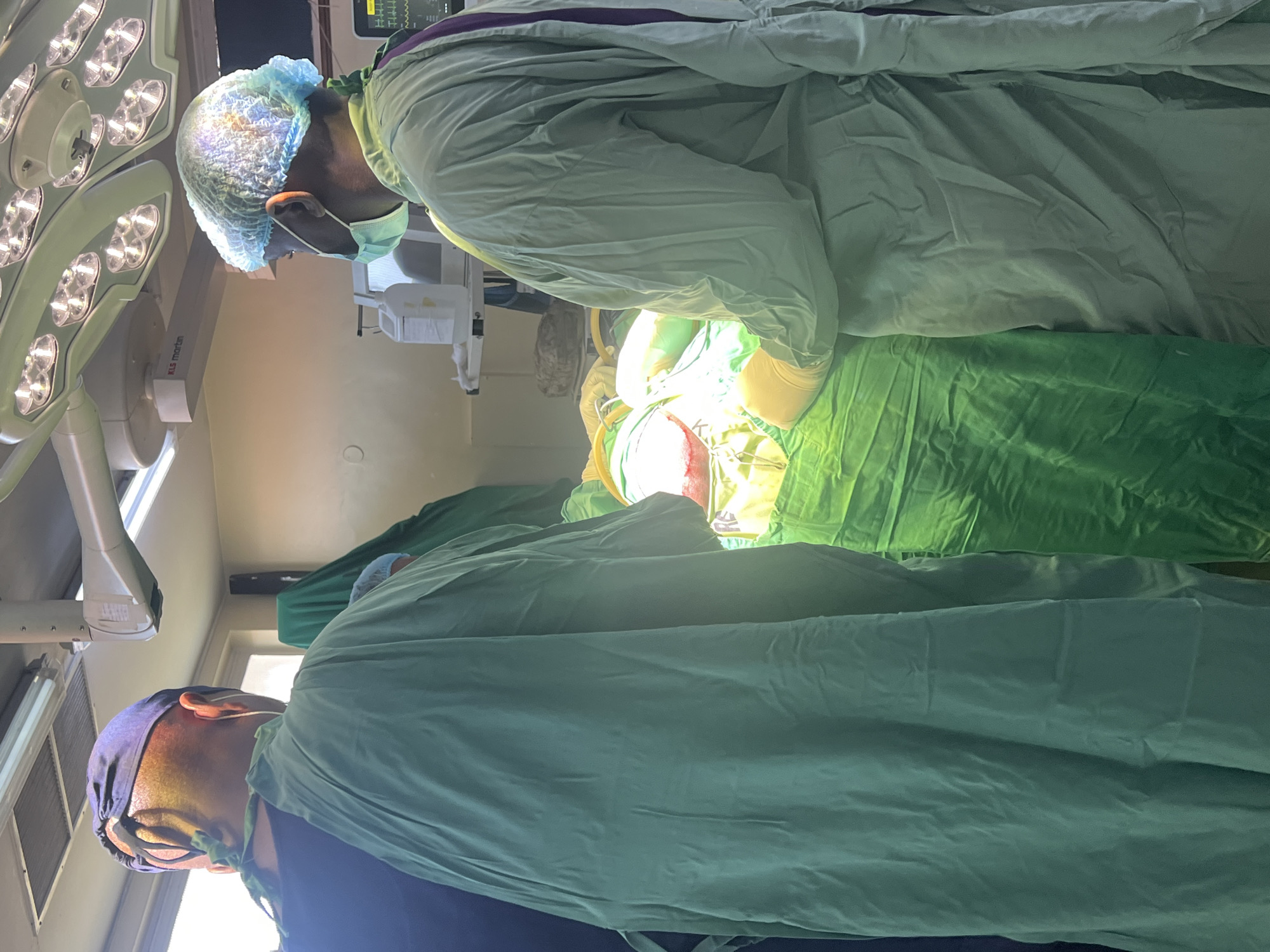 The image depicts an operating room scene, likely during a surgical procedure. Two individuals wearing surgical gowns, caps, and masks are visible. They are positioned around a patient covered with surgical drapes. Surgical lights are positioned above the operating area, illuminating the scene. Based on the file name and other available information, this image was taken in Nairobi, Kenya.