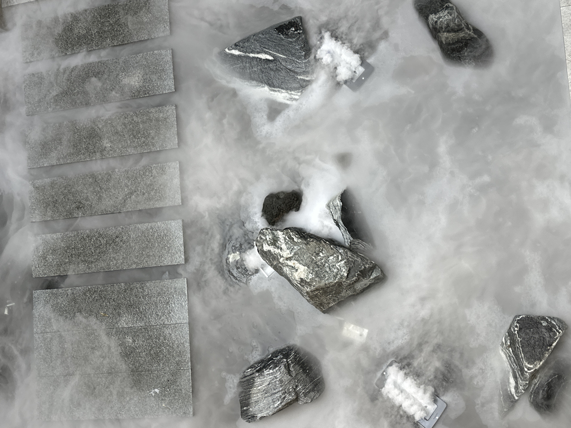An overhead perspective captures an artistic water feature or installation within a public or commercial space in Seoul, South Korea. The scene consists of a shallow body of water completely enveloped by dense, white, swirling vapor, consistent with a fog effect likely produced by dry ice. Multiple dark, irregularly shaped rocks and boulders are distributed throughout this misty expanse, some partially submerged. These rocks exhibit varied textures, with several displaying distinct white mineral veins or striations. Along the upper periphery of the frame, several rectangular, uniformly grey, textured stone slabs are arranged in linear formations, partially obscured by the pervasive fog. Dark-framed objects are observable within the water, from which streams of white mist are actively emanating, functioning as fog generators. No individuals are present in the scene.