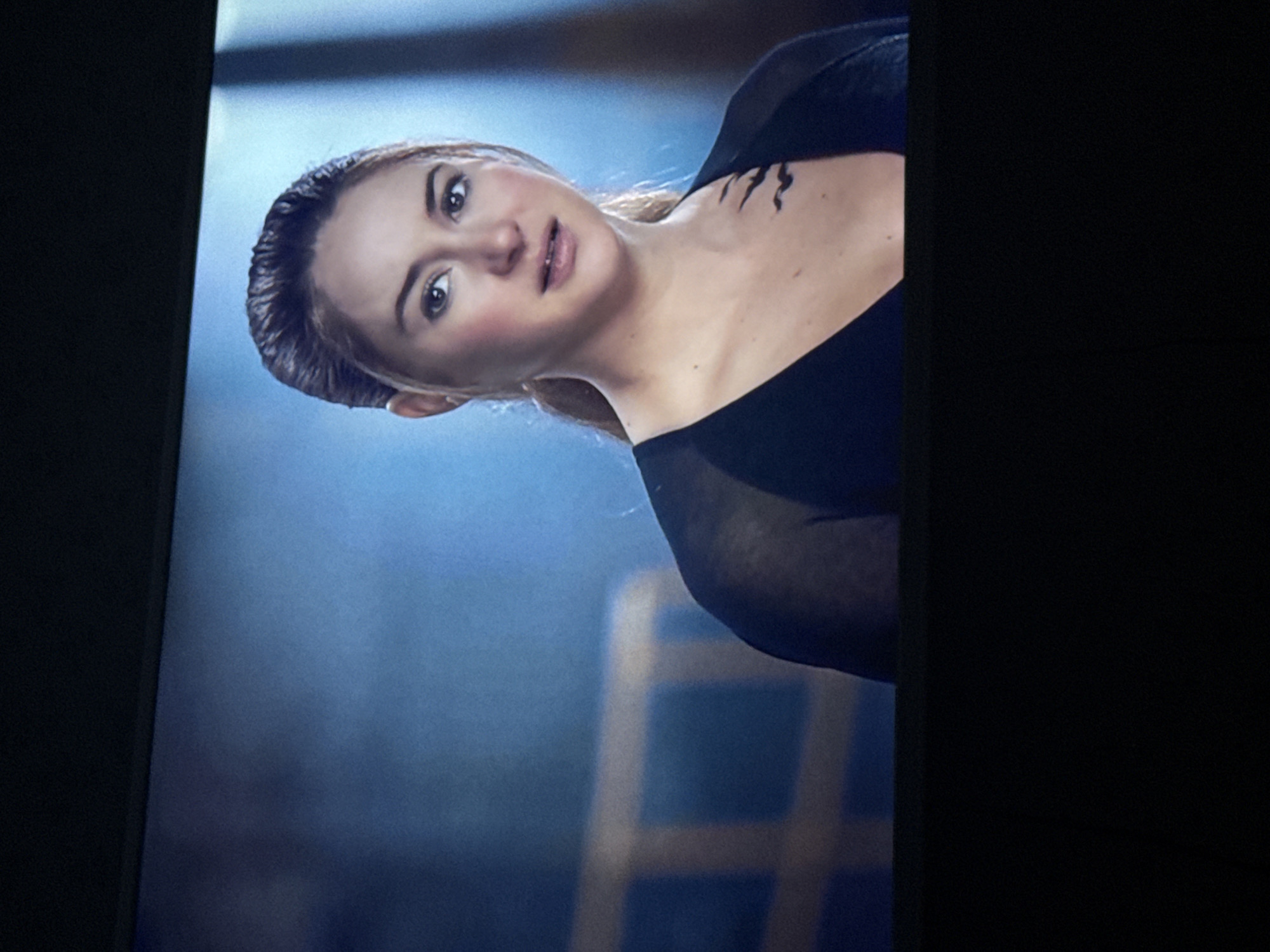 A medium shot depicts a woman with fair skin and light brown hair pulled back, wearing a black garment that reveals part of her shoulder and the top of her chest where a tattoo-like design is visible. She is looking towards the camera. The background features a hazy blue tone with a ladder-like structure slightly out of focus. There is no evident information to determine the location as Mytishchi, Russia.