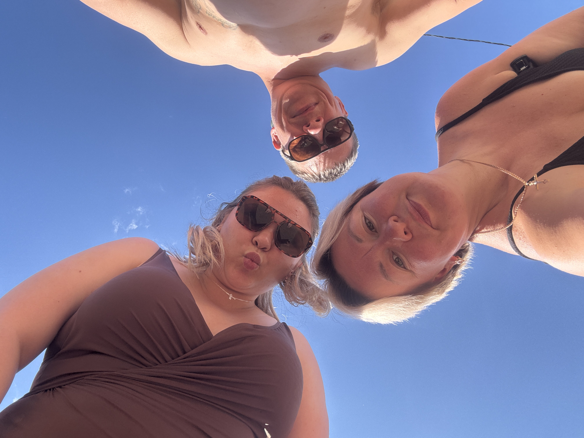 An extreme low-angle photograph captures three individuals looking downward into the camera against a clear blue sky, presumed to be in the city of Chaikovsky, Russia. In the foreground, at the bottom center, a middle-aged female with short, light blonde hair and a tanned complexion gazes directly at the lens with a neutral expression. She wears a black strap top and a gold chain necklace with a cross pendant; a single earring is visible in her right ear. Positioned to the upper left, a younger female with curly blonde hair and large dark sunglasses makes a puckering expression with her lips. She wears a dark brown sleeveless top featuring prominent ruching on the chest and a delicate necklace. To the upper right, a middle-aged male with short dark hair and dark sunglasses looks down, displaying a subtle smile. He is shirtless, exposing his torso, and a distinct scar or mark is visible on his upper right chest/shoulder area. A thin black wire or strap is visible near his right armpit. The background is a vibrant, cloudless blue sky, with a few faint wisps of white clouds visible in the upper left quadrant, indicating bright daylight conditions.