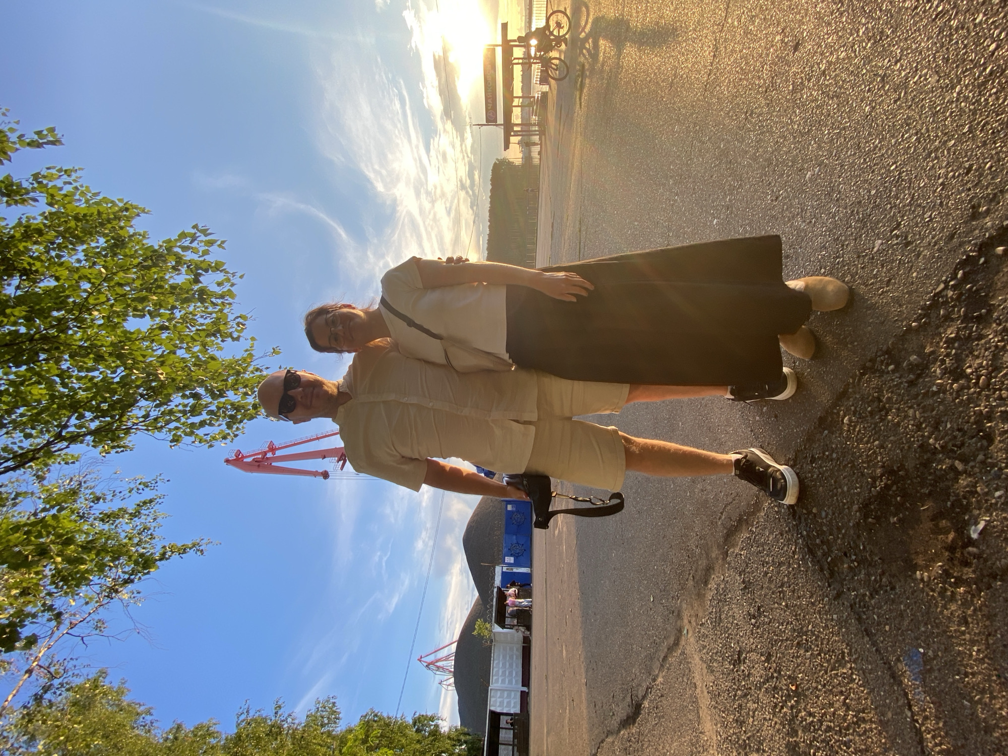 An outdoor, full-body, low-angle, natural light, eye-level photograph captures a couple standing side-by-side on an asphalt surface. The woman, on the right, is wearing glasses, a white short-sleeved t-shirt, and a long black skirt. The man, on the left, is wearing sunglasses, a light-colored short-sleeved button-up shirt, and beige shorts. He is holding a camera with a strap in his left hand. They are both facing the camera and appear to be smiling. In the background, a bright sky with some wispy clouds and the setting sun is visible. A large red crane is present in the mid-background, along with piles of dark material. To the right, a pier structure with the Cyrillic text "ЧАЙКОВСКИЙ" (Chaikovsky) is visible, and a person on a bicycle is silhouetted against the sun. The overall setting suggests a port or industrial area in the city of Chaikovsky, Russia, during sunset. Trees with green leaves hang into the frame from the top left and top right, casting shadows on the scene.
