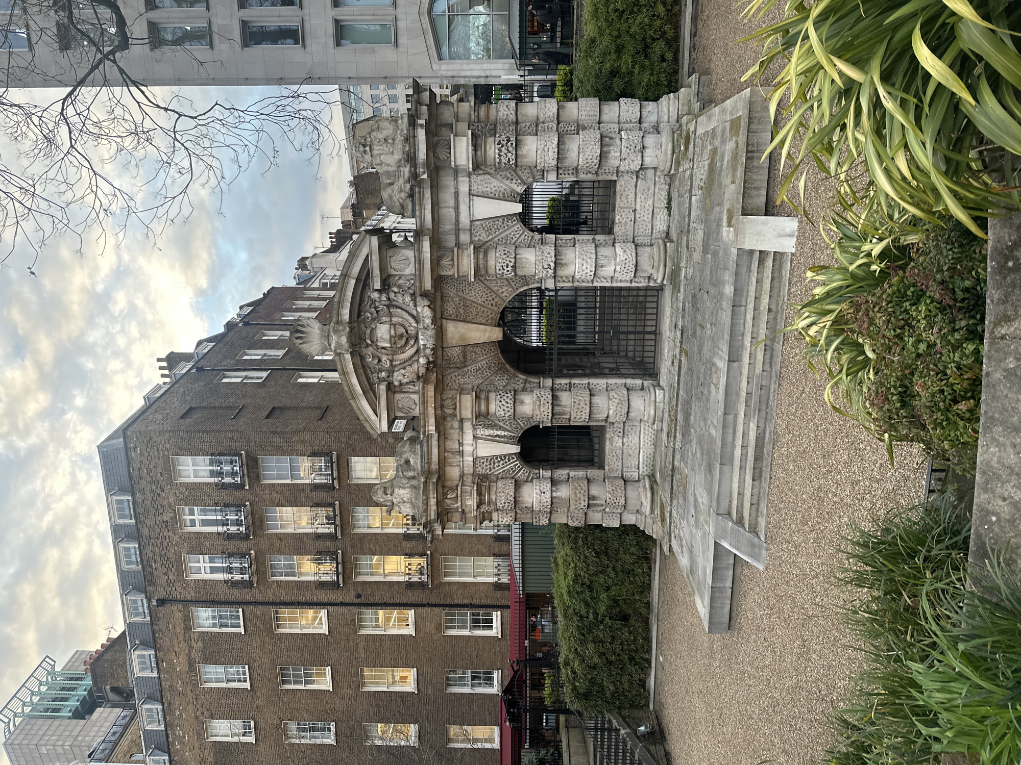 The image displays the Temple Bar, an ornate Baroque-style stone gateway, situated in Paternoster Square, City of London, United Kingdom. The gateway features three arched openings, the central one fitted with a black metal gate, supported by rusticated columns. Its upper section includes decorative carvings such as a coat of arms and is surmounted by two reclining lion sculptures. The structure stands on a raised stone platform, accessible by steps and a small ramp, which overlooks a large area of light brown gravel.
Surrounding the gateway are various buildings: to the left, a multi-story brown brick building, characteristic of 18th or 19th-century architecture, features numerous white sash windows, some with small black iron balconies, with interior lights active. A more modern building with a metallic rooftop structure rises behind it. To the right, a lighter-toned, contemporary building with large windows is visible, partially obscured by bare tree branches against an overcast sky. In the mid-ground, green bushes and tall grass-like plants are present. Indistinct human figures are visible in an outdoor cafe setting with red awnings to the left, behind a hedge, and a few dark-clad figures are partially observed to the right.