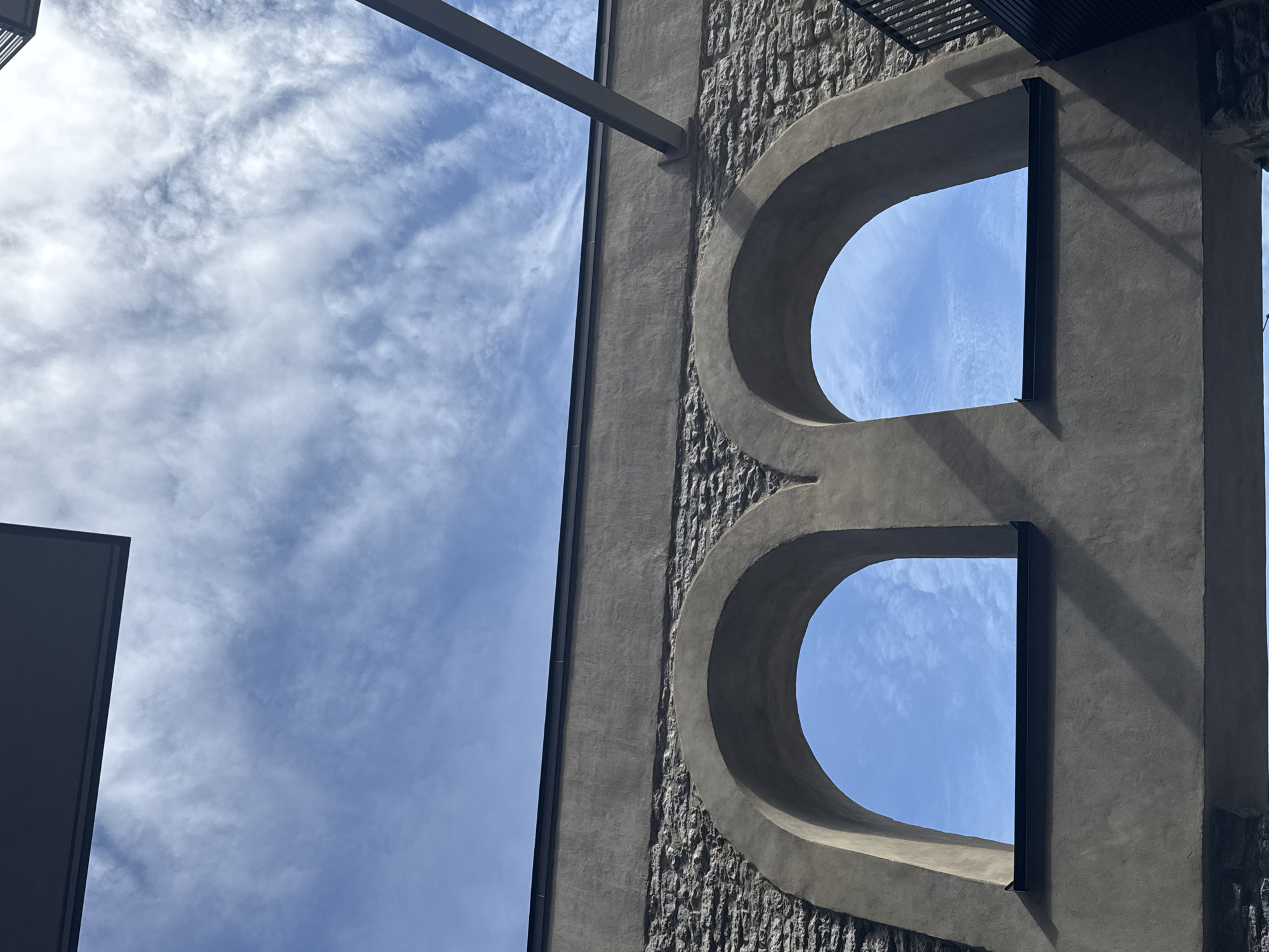 The image shows two arched openings in a stone wall, revealing a bright blue sky with scattered white clouds. The wall appears to be part of a building with a modern facade above the stonework. A dark, angled beam is visible in the upper right quadrant, intersecting the sky. The overall scene suggests an architectural detail set against a clear sky. This structure is located in the courtyard of the historic Rotermann Quarter in Tallinn, Estonia.