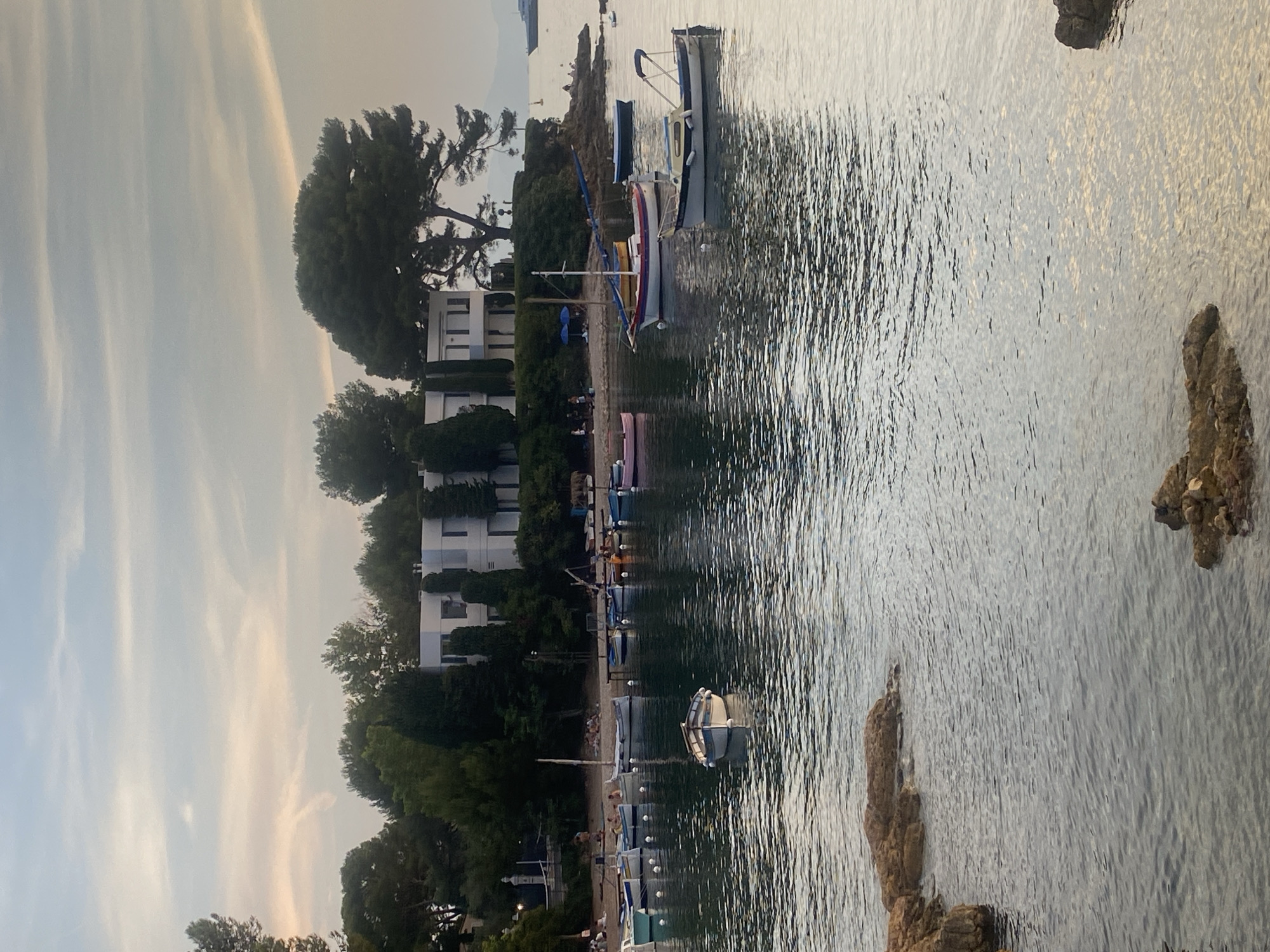 The image depicts a tranquil harbor scene in Antibes, France, likely at sunset. In the foreground, gentle waves lap against rocky shorelines on the left and right sides of the frame. The middle ground is filled with a collection of various boats, some moored and others gently bobbing on the water. One small, traditional wooden boat with a white hull and blue trim sits prominently in the center. Further back, a line of more modern-looking vessels is anchored along a sandy beach.
On the beach, several people are visible, some sitting, others standing or walking. Two blue umbrellas offer shade. Behind the beach, a white, modern villa with large windows and a flat roof is nestled amongst lush green trees, including tall cypress trees and a large pine tree with distinctive, sprawling branches. The sky above is a pale blue, streaked with soft, hazy clouds tinged with warm sunset colors. In the far distance, a large ship is visible on the horizon. The overall atmosphere is peaceful and serene.
