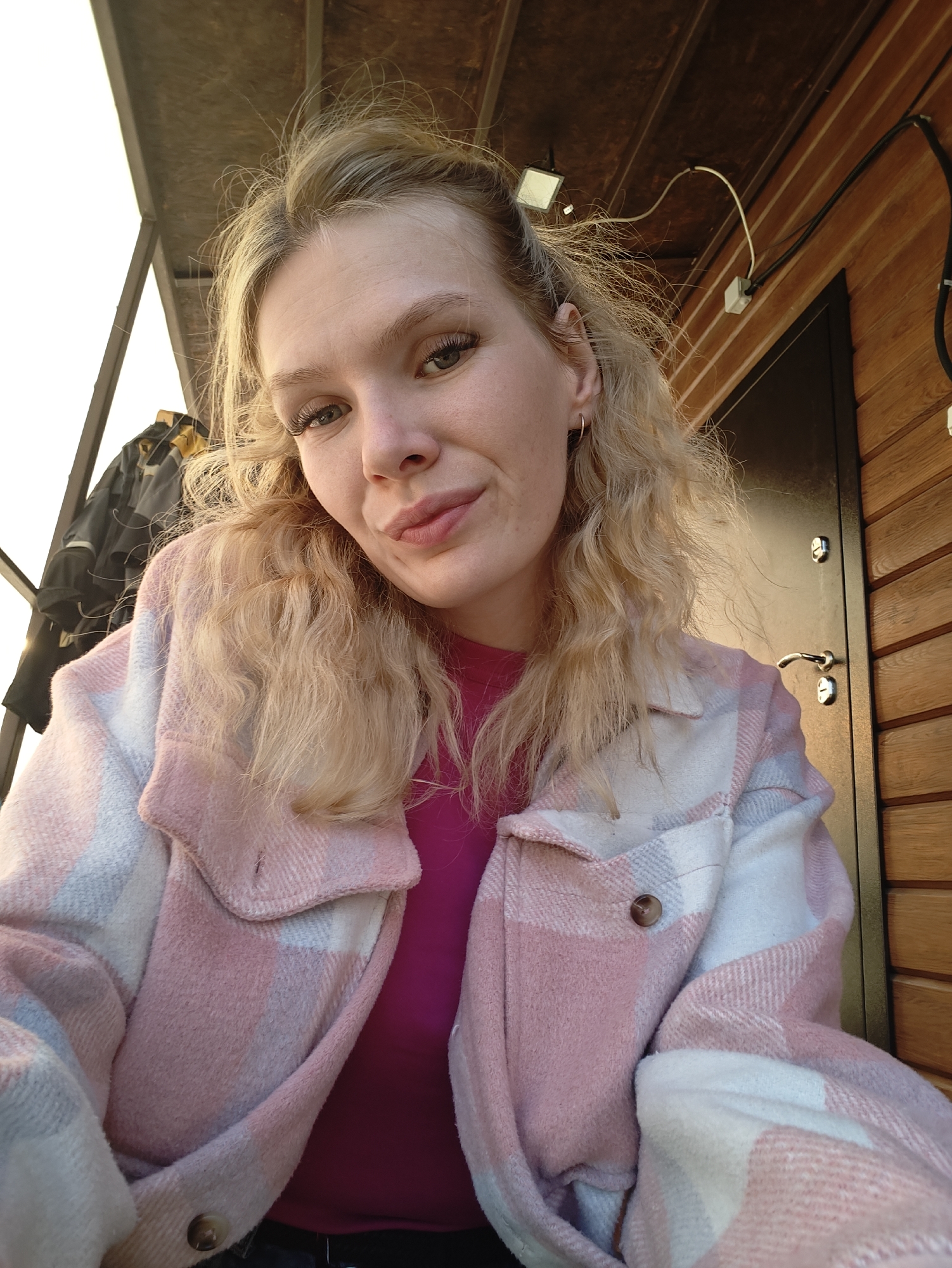A close-up, selfie-style, eye-level shot shows a young Caucasian woman with blonde hair pulled back and appearing windswept. She has green eyes and is wearing a pink and white plaid jacket over a magenta top. She is looking at the camera with her lips slightly pursed and a soft smile. The background consists of a wooden wall with vertical planks, a dark door with a silver handle, and some electrical wires and fixtures. Above, a wooden ceiling with visible beams is present, and to the left, a section of a white structure and a dark garment on a hanger are visible. The lighting suggests an outdoor setting during daylight. The location is identified as Russkiy Vozhoy, Russia.