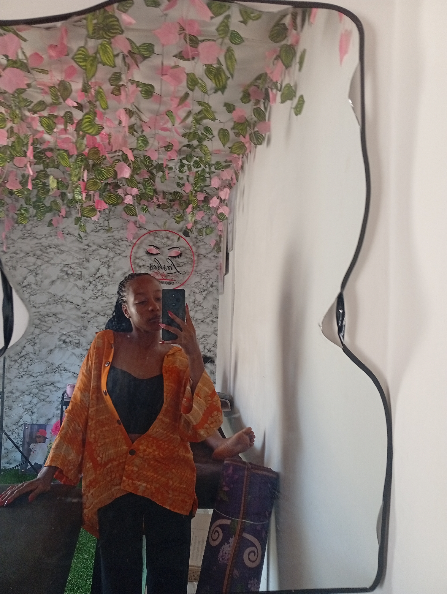 A young woman stands in front of an irregular-shaped mirror in a room decorated with pink and green floral garlands hanging from the ceiling. The woman, who appears to be taking a selfie, is wearing a black top, an orange patterned shirt, and black pants. A logo featuring stylized eyelashes and the word "Lashes" is visible on the wall behind her. A glimpse of a foot can be seen reflected in the mirror on what appears to be a purple mat, along with other objects like a candle, a picture, and a light. The room seems to be in a commercial or personal space, possibly a beauty salon, likely located in Narayi, Nigeria.