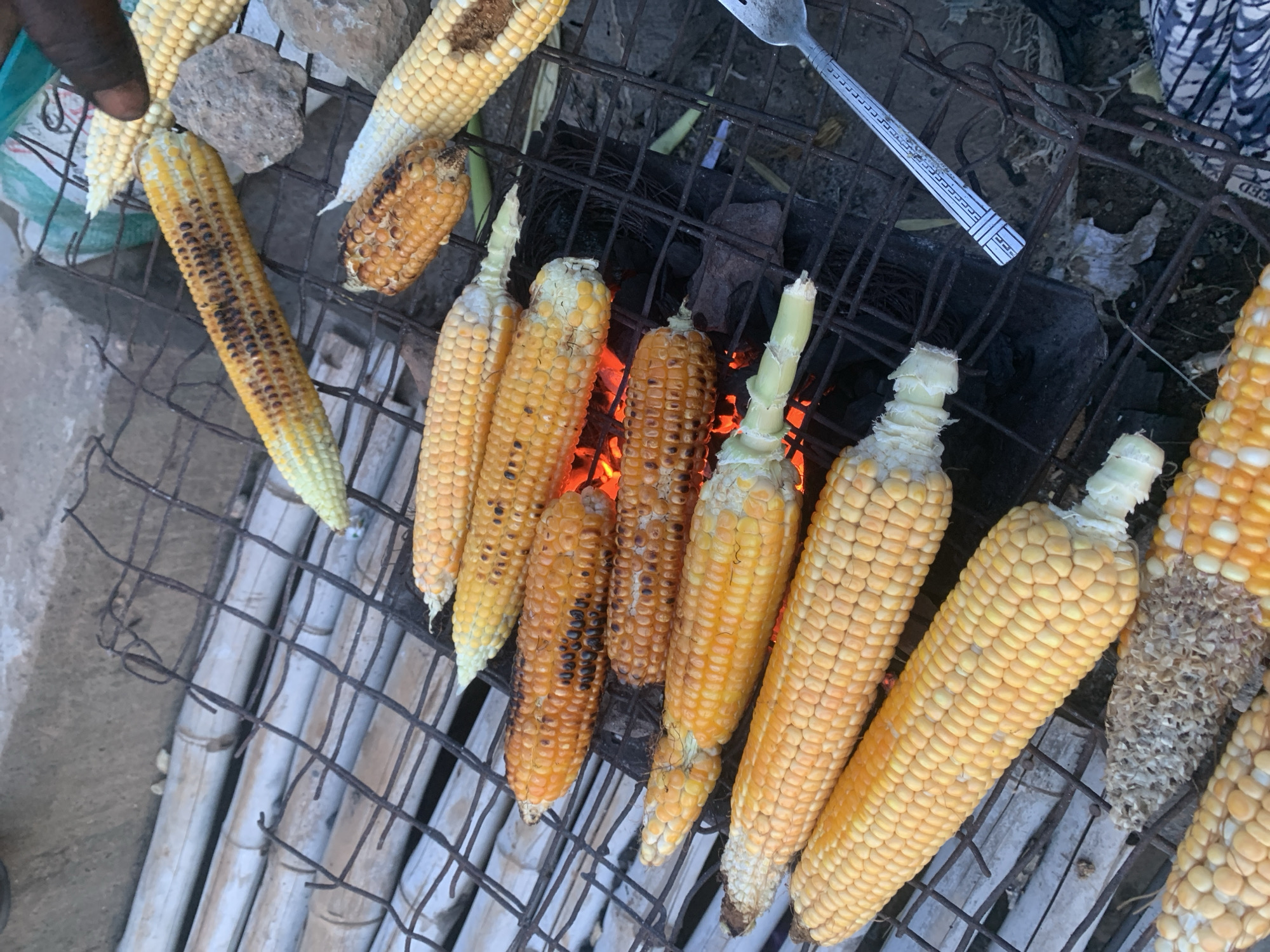 The image shows an outdoor cooking scene where several ears of corn are being grilled over hot coals. The corn cobs are arranged on a metal grill placed over glowing red embers. Some of the corn appears to be partially roasted with some charred kernels. The background consists of a bamboo-like structure and a rough surface that could be earth or concrete. A hand is visible in the upper right corner of the image, reaching towards the corn. A fork or spoon is also present on the right side of the grill. The overall setting suggests a casual, possibly street-side, food preparation environment. The specific location in Keffi, Nigeria, is not visually identifiable from the image itself.