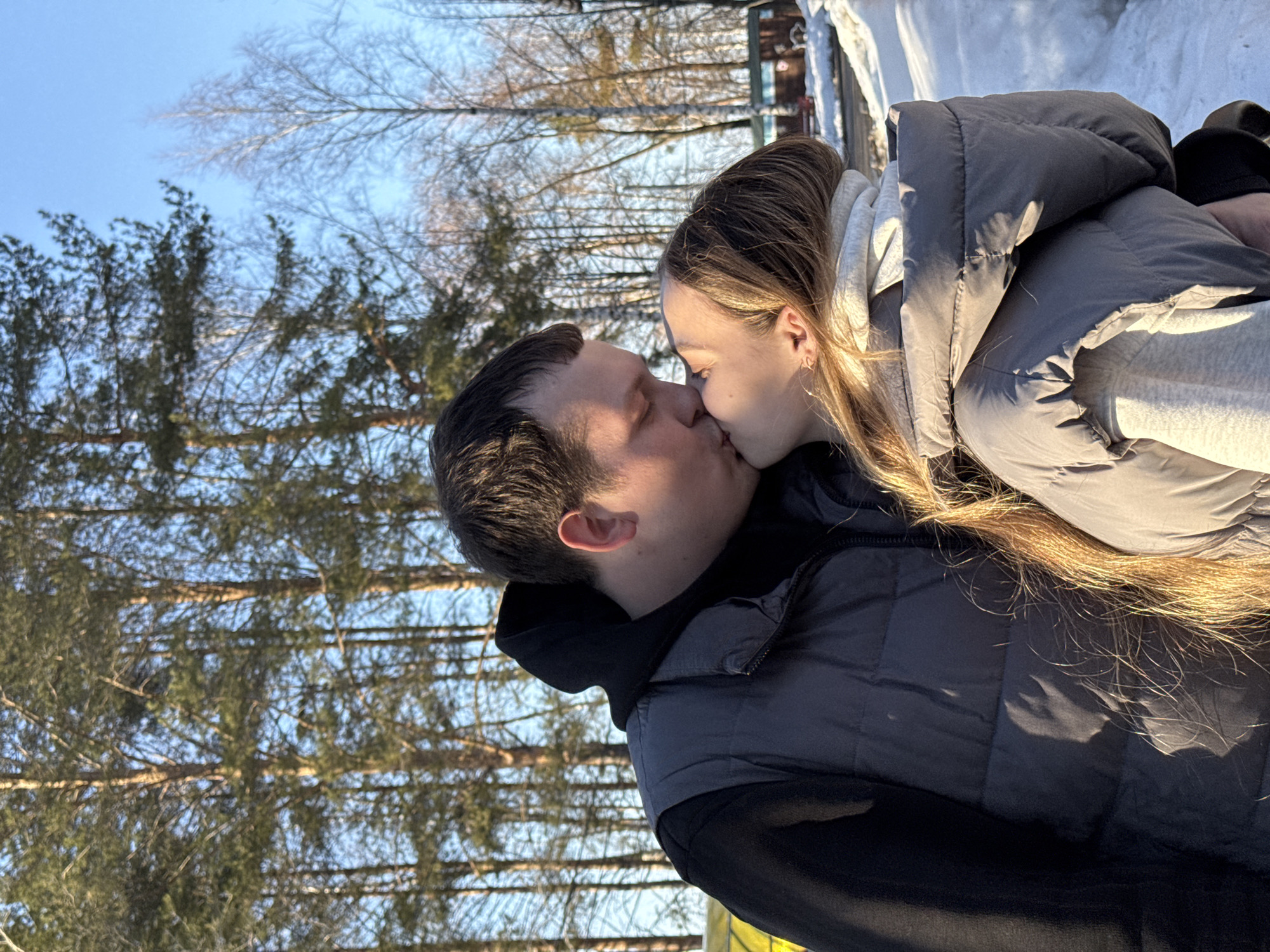 The image shows a man and a woman kissing outdoors in Chaikovsky, Russia. The background features trees and snow, suggesting a park or wooded area. The sky is blue. The woman has long, light brown hair and is wearing a grey puffer jacket with a light grey hoodie underneath. The man is wearing a dark puffer vest over a black hooded sweatshirt.