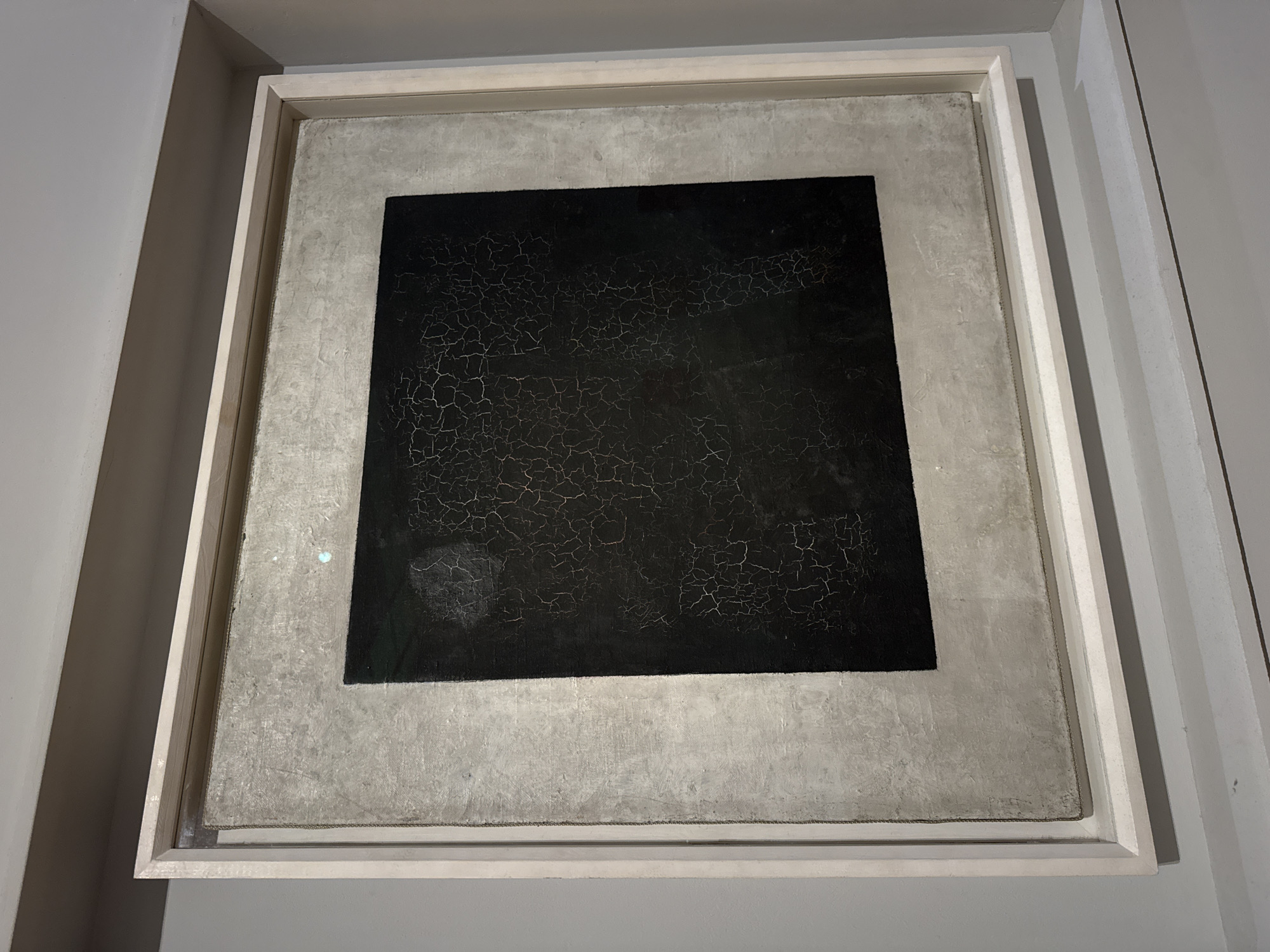 The image shows Kazimir Malevich's "Black Square" painting, a famous example of Suprematism, on display. The artwork is a black square on a white background, framed with a light-colored frame. The painting is displayed on a gray wall. The location is likely a museum in Moscow, Russia, where the Tretyakov Gallery houses a version of this iconic work. The image focuses on the artwork itself and its presentation within the gallery setting.