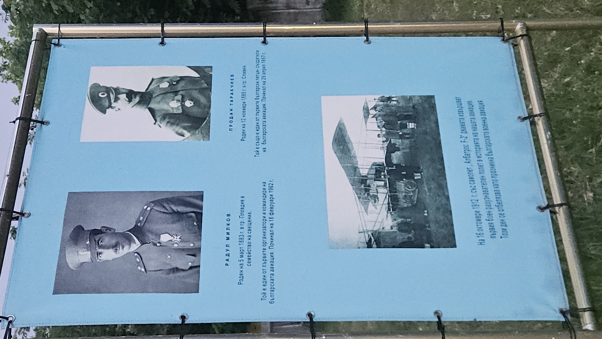 The image shows a blue informational sign with photographs and text in Bulgarian. On the left side are two headshots of men in military uniforms, identified as Radul Milkov and Prodan Tarachiev. The text below the pictures states that Milkov was born on March 5, 1883, in Plovdiv, and was one of the first organizers and commanders of Bulgarian aviation, dying on February 16, 1962. The text about Tarachiev indicates he was born on November 12, 1885, in Sliven and was also one of the first Bulgarian pilots, dying on April 29, 1957. To the right of the portraits is a black and white photo of an Albatros F-2 airplane surrounded by people. The text below explains that on October 16, 1912, these two pilots flew the plane in a reconnaissance mission. The sign is suspended by loops to a metal frame. The background shows greenery, suggesting the sign is in a park or outdoor area.