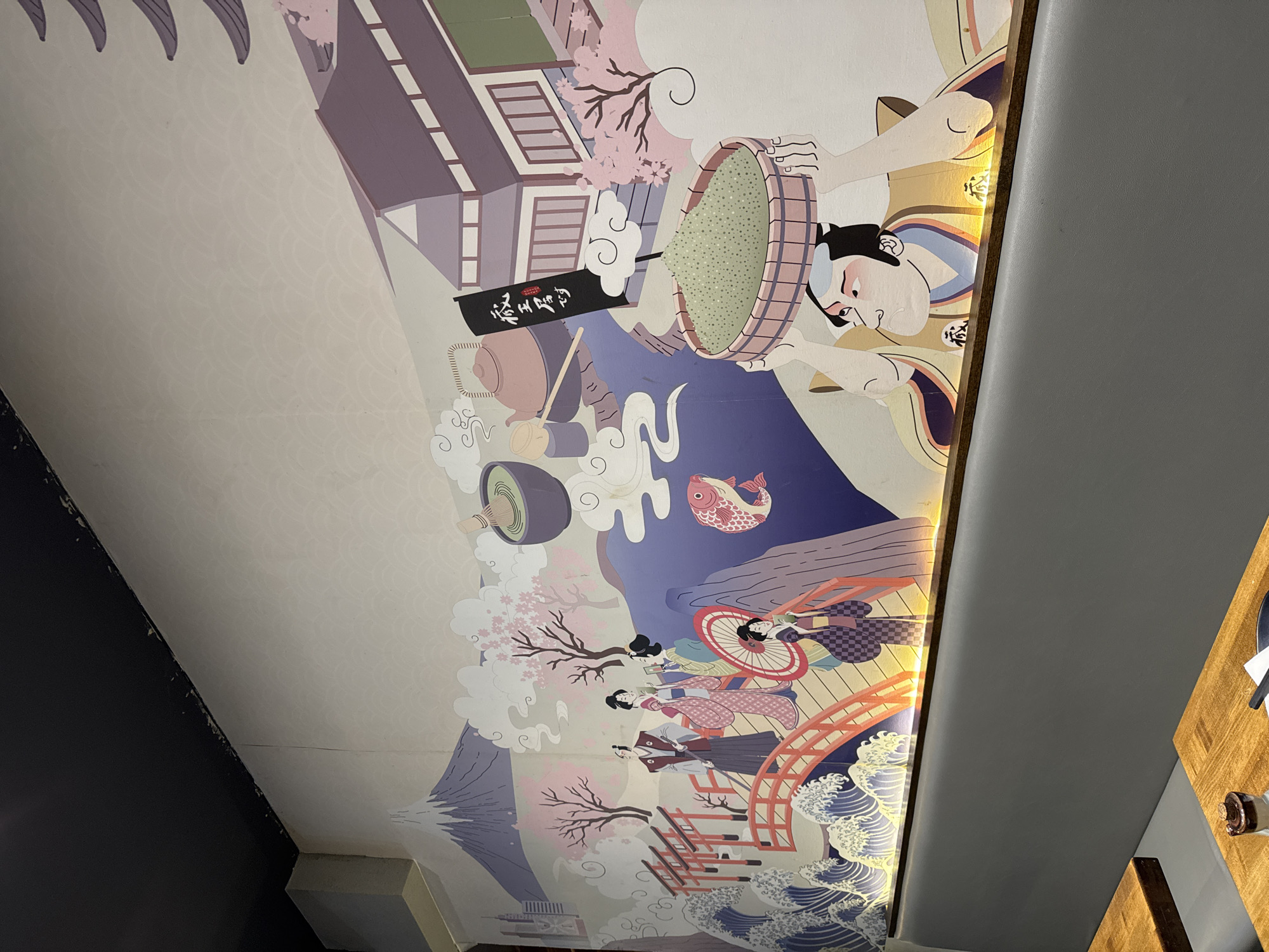 This is a photograph of a mural painted on a wall in what appears to be a restaurant. The mural depicts a Japanese-themed scene with Mount Fuji in the background, cherry blossoms, traditional Japanese architecture, and figures dressed in kimonos. There are also elements related to tea culture, such as a bowl of matcha and a teapot. The mural is illuminated by yellow lights running along its base. The bottom of the image shows a grey upholstered bench seat and wooden tables, consistent with a dining establishment. The style is illustrative, with flat colors and bold outlines. The mural is a vibrant and detailed artwork designed to evoke a sense of Japanese culture. The location is not specified in the image.