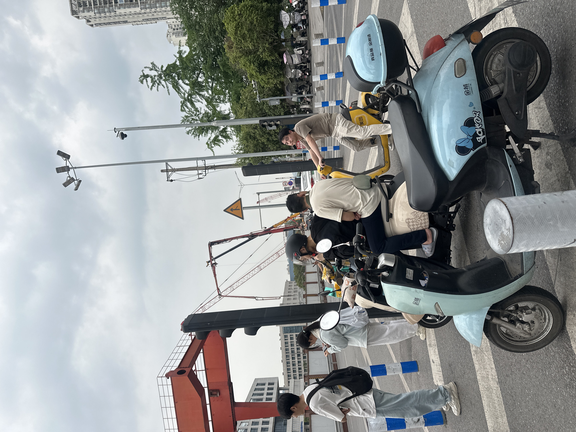 The image, taken in Chongqing High-tech Zone, China, shows several people standing on a crosswalk next to a row of parked electric scooters. The scooters are a mix of blue and yellow, some with storage compartments. People are focused on their phones. Construction cranes and buildings are visible in the background. The sky is overcast. Streetlights and traffic signals are present.
