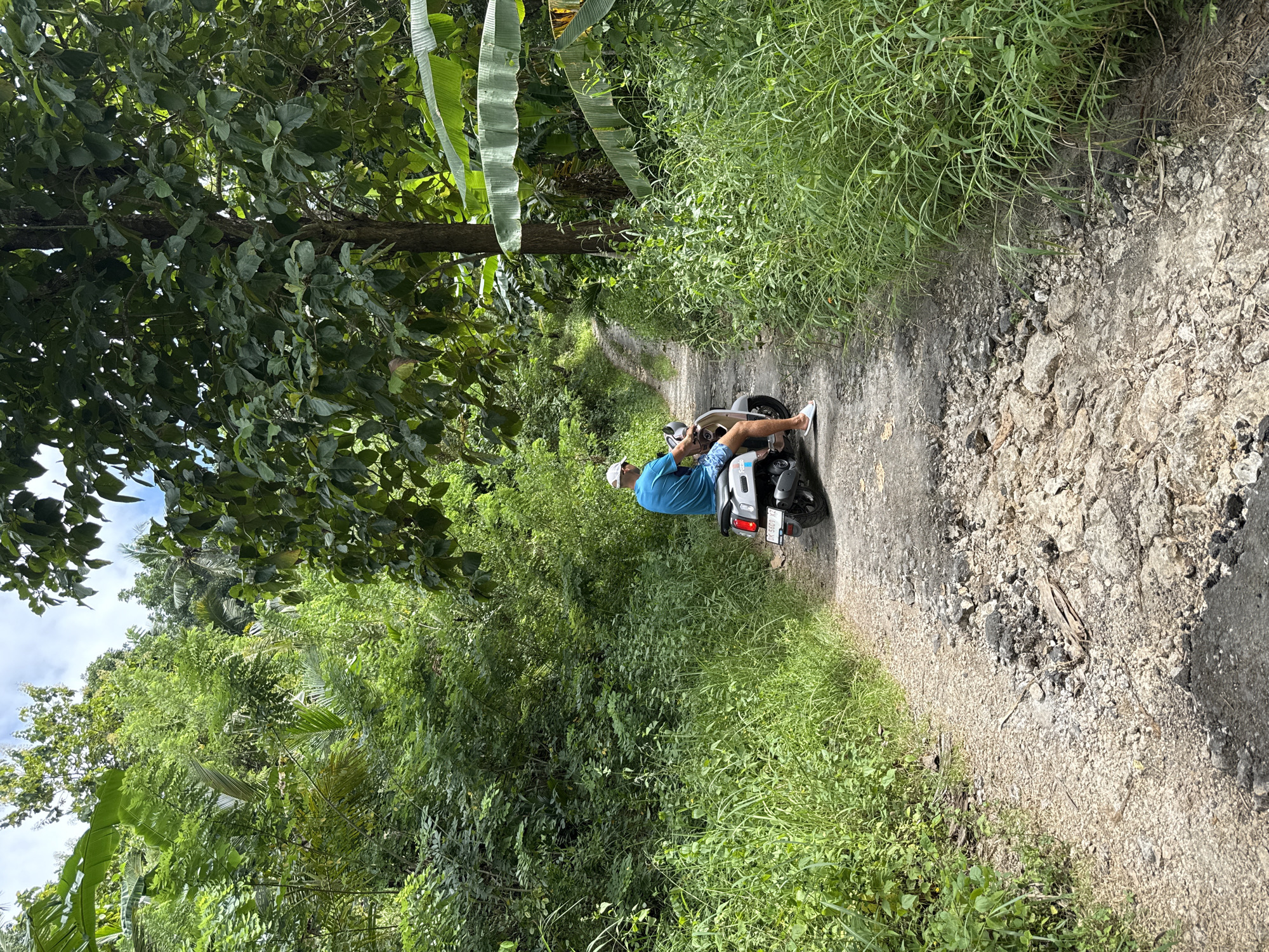An outdoor scene in Tanglad, Indonesia shows a man riding a scooter down a narrow, unpaved road flanked by dense vegetation. The road surface is uneven, composed of dirt, gravel, and asphalt fragments. On either side, lush greenery including trees, shrubs, and tall grasses encroach upon the path. The man, wearing a blue shirt and white hat, is the primary subject. He is operating a white scooter with visible license plate number 'M 5609 FG'. He is facing away from the camera. The overall environment is rural and overgrown, typical of a tropical Indonesian landscape. The image is shot from a slightly elevated perspective.
