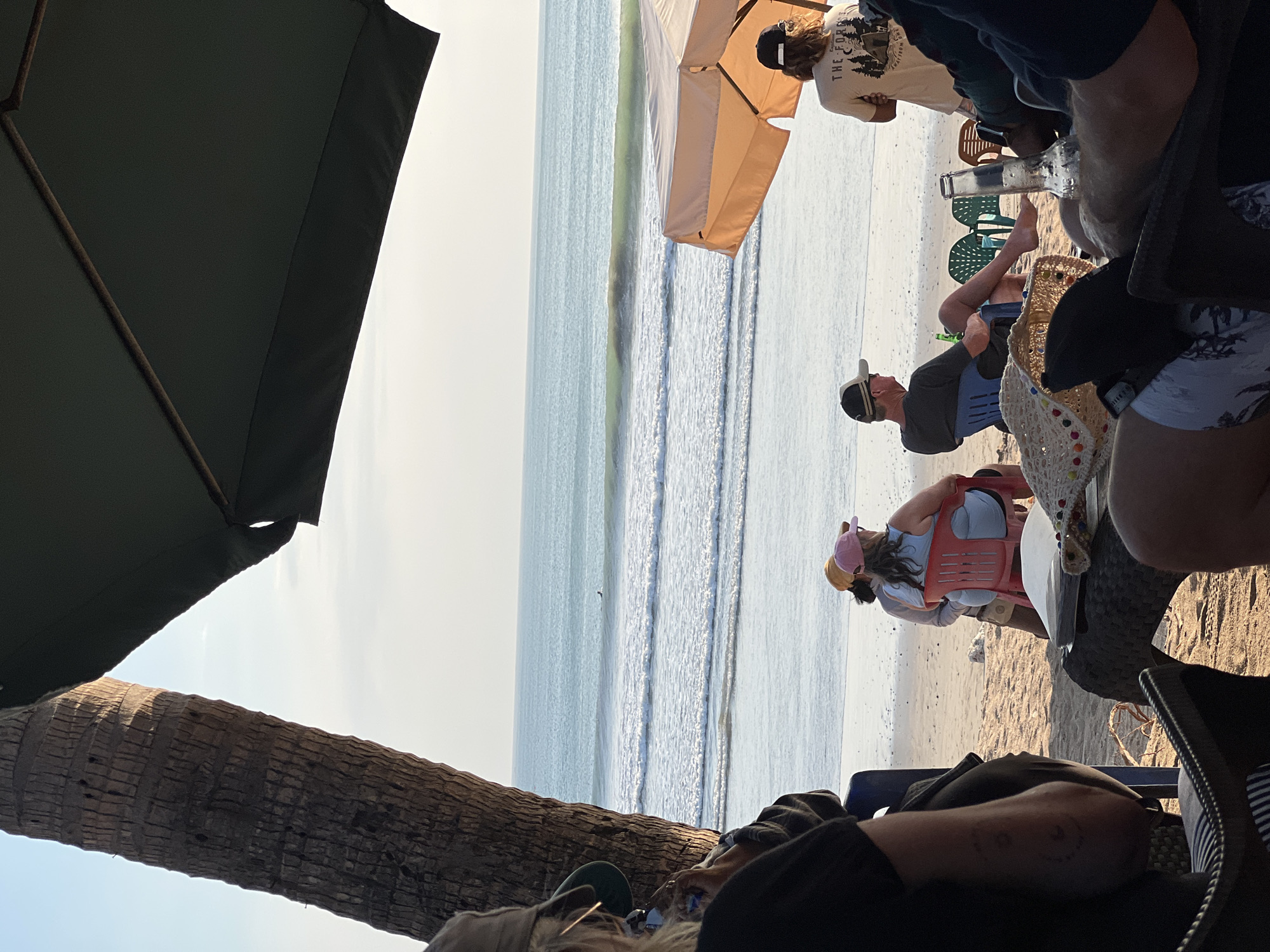 The image depicts a beachfront scene in Kuta, Indonesia, likely in the late afternoon based on the lighting. A group of people are seated on a sandy beach, some under the shade of umbrellas. In the foreground, individuals are seated at tables, with a straw hat and an empty glass bottle visible on one table. To the left, the trunk of a palm tree is prominent.
Beyond the immediate foreground, several people are seated on chairs facing the ocean. One woman in a red chair wears a blue top and a pink cap. A man in a blue chair wears a cap and a dark shirt, with a green bottle near him.
The ocean stretches towards the horizon, with gentle waves breaking on the shore. A lone surfer can be seen in the distance. The sky is clear and a light blue, with the sun casting a warm glow on the scene. A large, dark umbrella partially covers the upper right portion of the image. Another lighter-colored umbrella is visible to the right, behind some of the seated individuals. The atmosphere suggests a relaxed, casual setting at a beachside establishment.