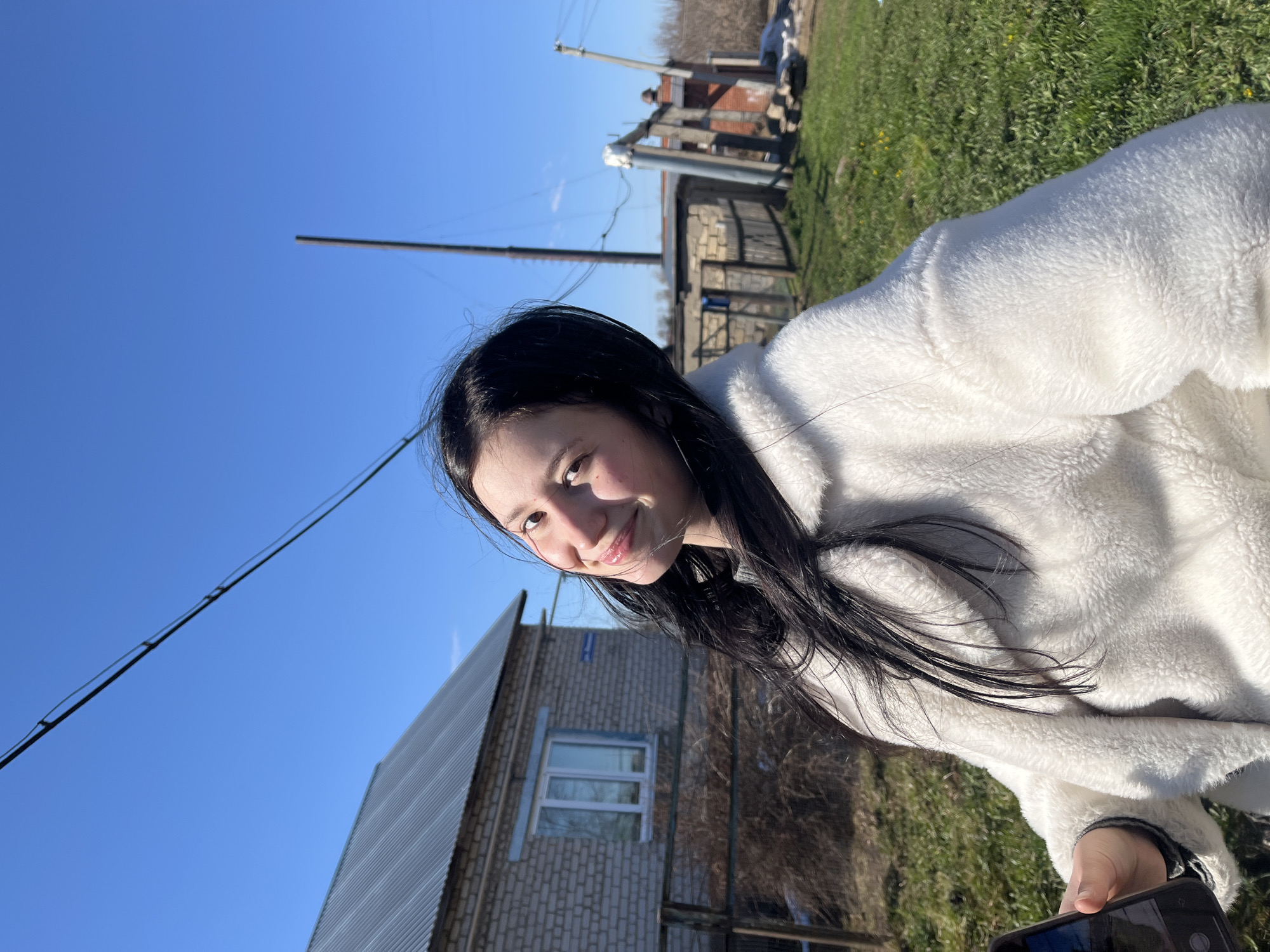 A young woman with black hair is smiling at the camera. She is wearing a white, fluffy jacket. The background includes a blue sky, a brick building with a gray roof, grass, and a utility pole with wires. In the distance, there is another building of brick and wood. Based on the landscape and architectural styles, the location could be in the city of Фоки, Russia. She appears to be holding a cell phone, presumably taking a photo of herself.