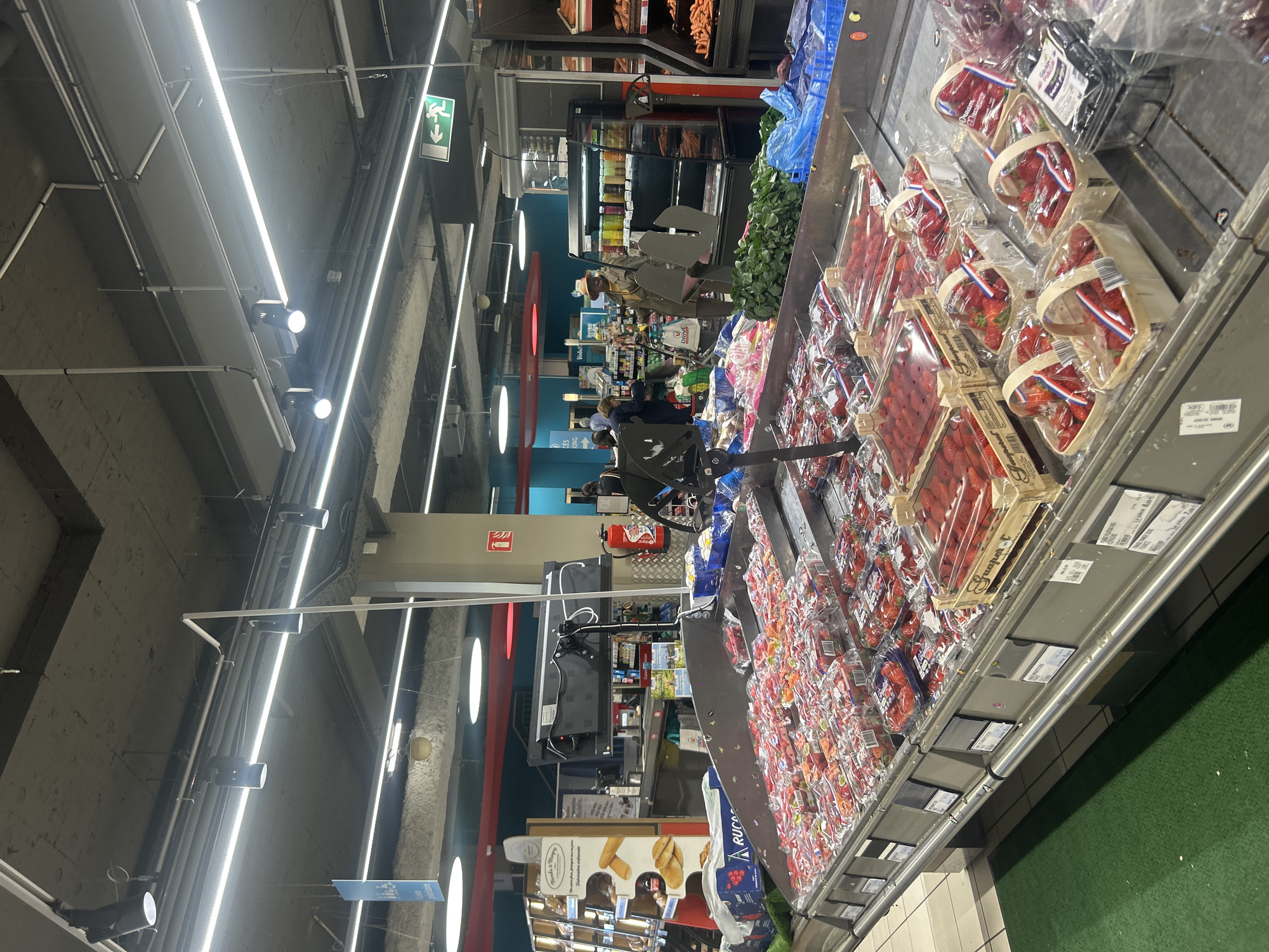 This image captures an indoor scene within a modern supermarket or grocery store, likely in Noisy-le-Grand, France, given the visible French text. The primary focus is a large produce display, predominantly featuring numerous packaged strawberries.
The scene is brightly lit by linear fluorescent lights and spotlights mounted on the high, concrete ceiling, which also features exposed pipes and wiring. In the foreground, dozens of plastic containers and wooden crates filled with strawberries are meticulously arranged on a low display unit. Some containers bear labels such as "Dream Manann's" and "Guapert FRANCE". Green artificial turf flooring is visible directly adjacent to the produce display, transitioning into grey tiled flooring further into the store.
In the mid-ground and background, several shoppers are visible, some pushing shopping carts, browsing other aisles. One individual with a distinctive hat and brown attire is visible near other produce displays. Refrigerated display cases with beverages and other products line the walls. To the left, a display stand for "Vinted" and a bakery section sign featuring "agricole & Mage officielle" with images of baguettes are discernible.
Notable details include a green exit sign with a downward-pointing arrow and a pictogram of a running person mounted on the ceiling. A red fire extinguisher and its corresponding sign are also visible on a wall. A black monitor or display screen is positioned on a stand near the center, possibly for self-checkout or informational purposes. The overall impression is a typical, bustling retail environment during an indeterminate time of day. Other visible text includes "ARUCA," "CES KING," and "DRIVE."