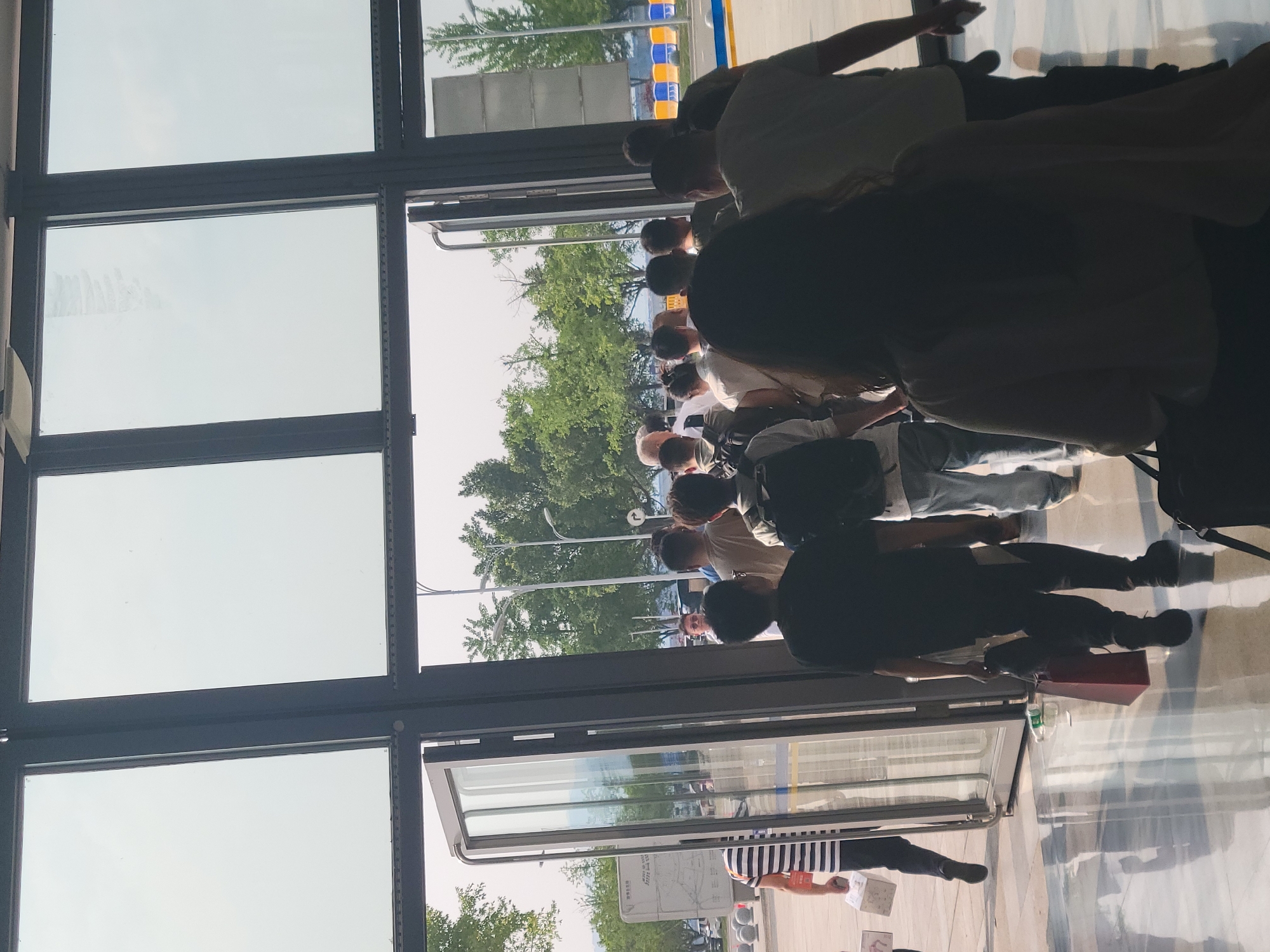 A photograph taken from inside a building looking outward through a glass doorway. A large group of people stand outside, seemingly waiting in a line. Trees and greenery can be seen in the background along with streetlights. A bus stop is visible to the left of the group. The location is Liangjiang, China.