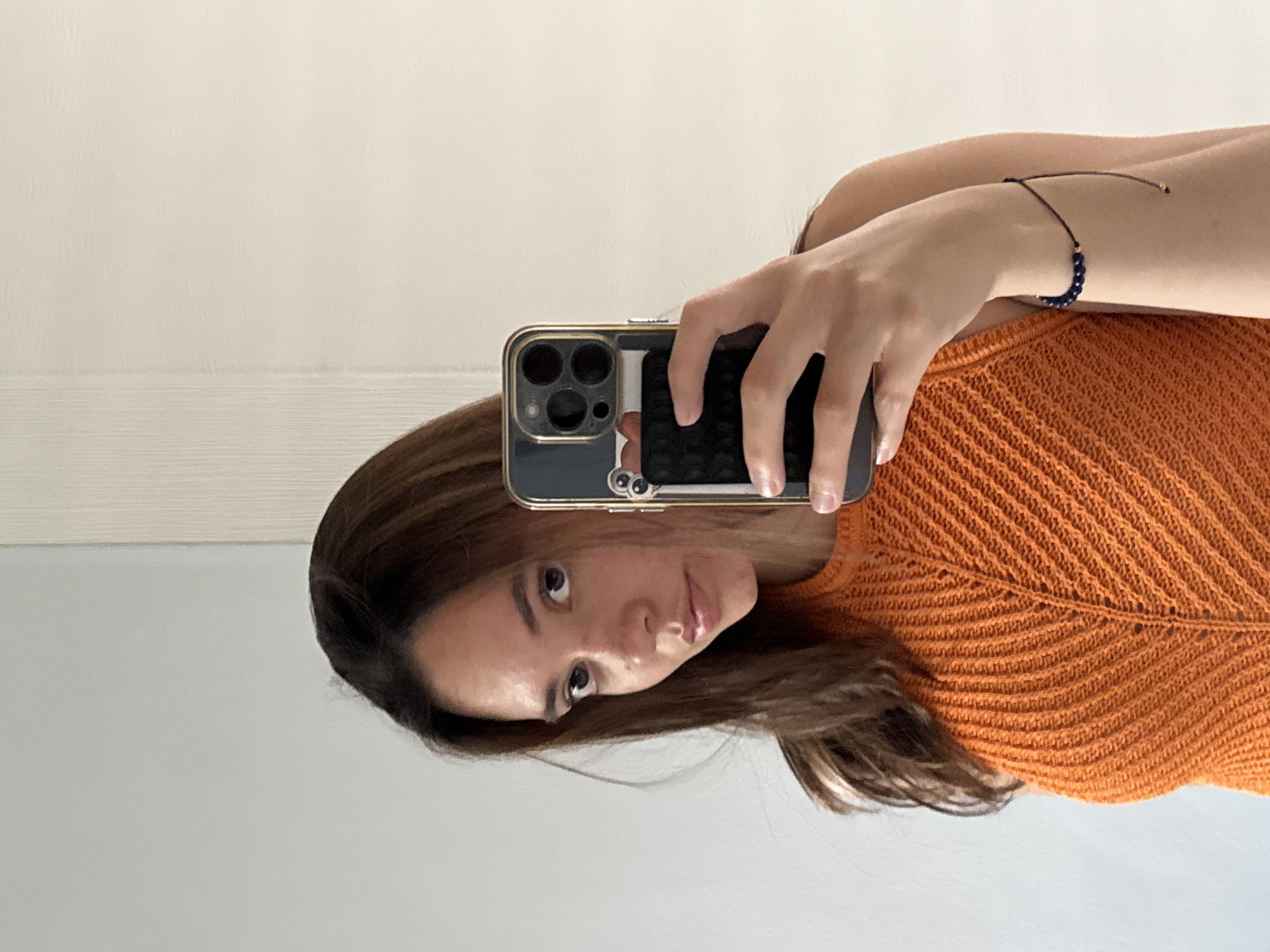 A woman is taking a selfie indoors. She holds a dark-colored phone with a gold-toned case in her right hand. Her hair is brown and shoulder-length. She is wearing an orange knitted top. A bracelet with dark blue beads is visible on her right wrist. The background consists of two tones, cream and light gray. The location is not discernible from the image.