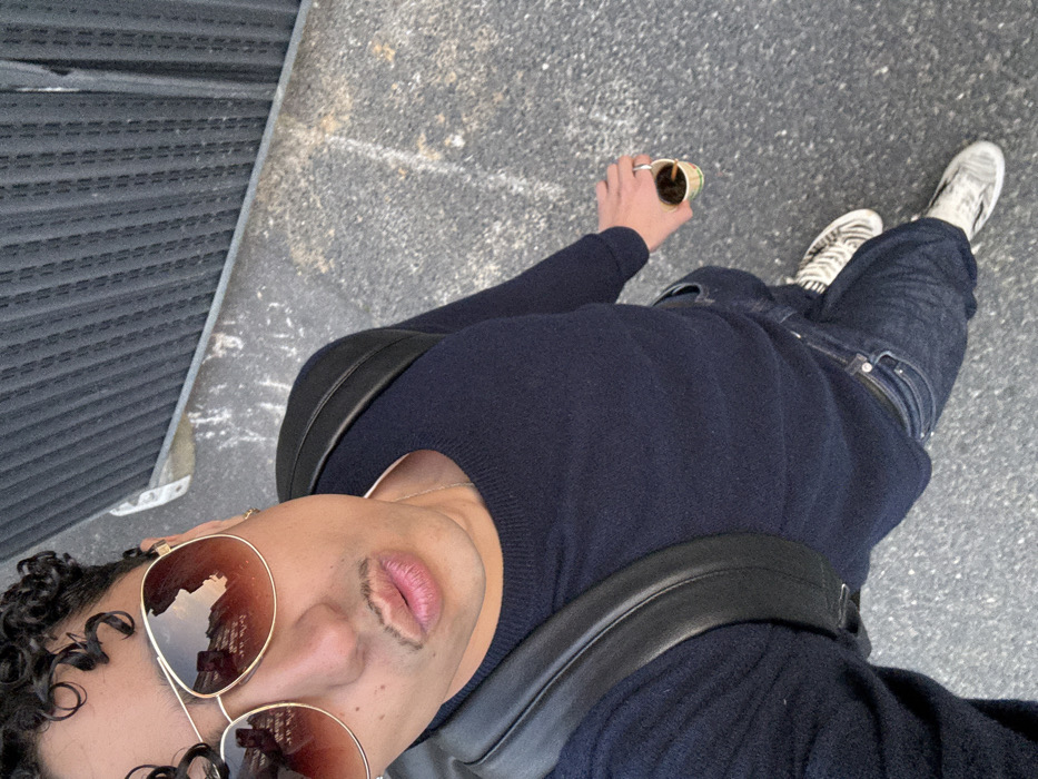 A high-angle selfie captures a male individual with curly dark hair, wearing aviator-style sunglasses and a styled mustache. He is dressed in a dark navy blue long-sleeved sweater, dark blue jeans, and white/grey patterned slip-on sneakers. A black leather backpack is visible on his shoulders. He wears a gold-colored earring in his right ear and a silver-colored ring on his left ring finger. His lips are pursed. In his left hand, he holds a small, multi-colored disposable paper cup containing a dark liquid. The individual stands on dark grey asphalt pavement adjacent to a dark, vertically corrugated wall or shutter. Reflections in the sunglasses reveal light-colored buildings with distinct windows and balconies, consistent with the architectural style prevalent in Paris, France.