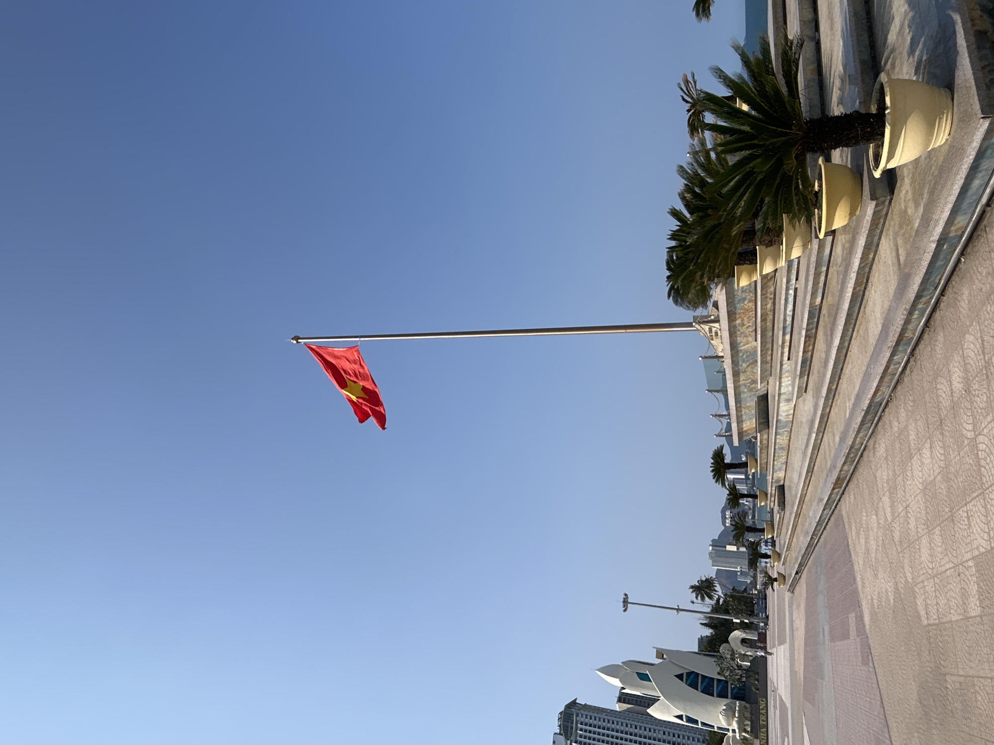 The image shows a clear blue sky above Nha Trang, Vietnam. A Vietnamese flag waves prominently on a metal pole. The right side of the image captures a section of a seaside promenade featuring potted palm trees and a paved walkway. In the distance, buildings and streetlights are visible, including a white architectural structure with the city's name etched on it.