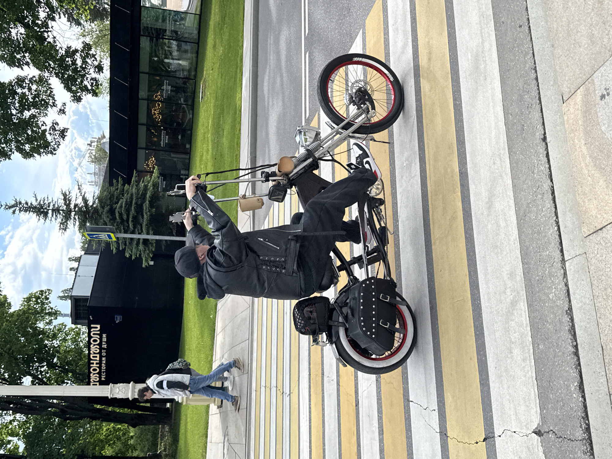 A man is riding a custom chopper-style bicycle across a pedestrian crosswalk. He is wearing a black leather jacket, black pants, and black and white sneakers. A black cap covers his hair. The bike is black with white-walled tires and a red accent stripe. A black leather bag is attached to the back of the bike. In the background, a building with the name "FONDYUGOLI" in Cyrillic (ФОНДЮГОЛИ) is visible. This is likely a restaurant, and the phrase underneath translates to "Restaurant from the soul". A person is walking on the sidewalk in front of the building. The photo was taken in Moscow, Russia.