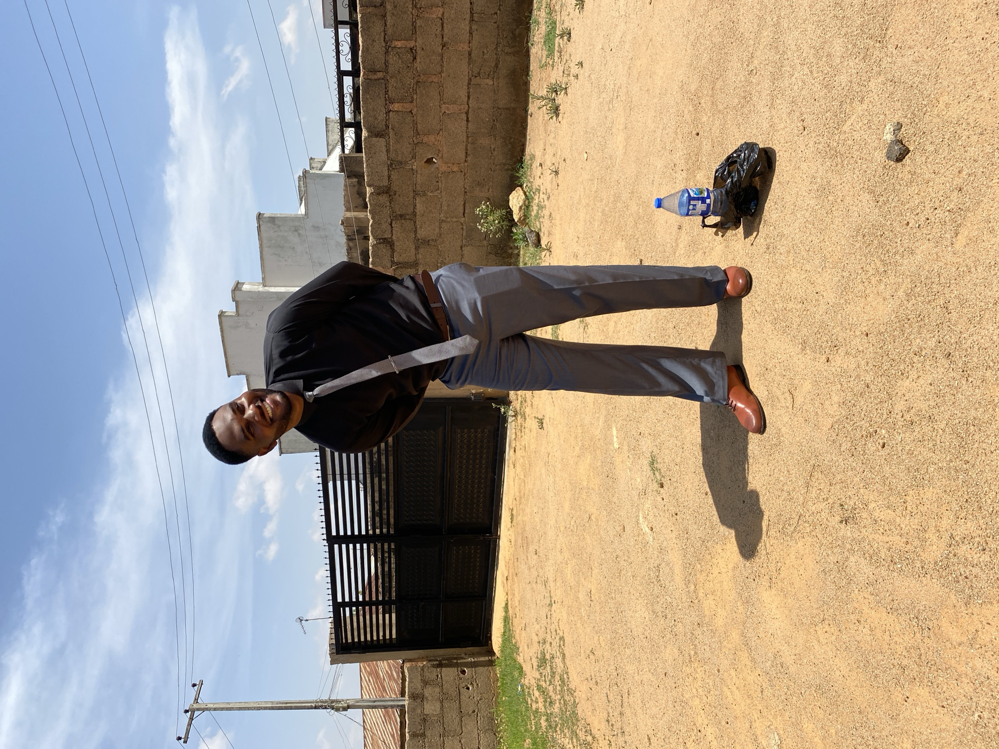 A man stands on a sandy surface outdoors. He wears a black long-sleeved shirt, grey trousers, and brown leather shoes. A brown leather belt is visible. He is smiling and looking at the camera. Behind him is a dark metal gate, and beyond that, a white building is partially visible. To the right, there's a rough brick wall. On the ground, there is a plastic water bottle inside of a black plastic bag and a small rock. The background includes a clear blue sky with light clouds. The location is Bukuru, Nigeria.