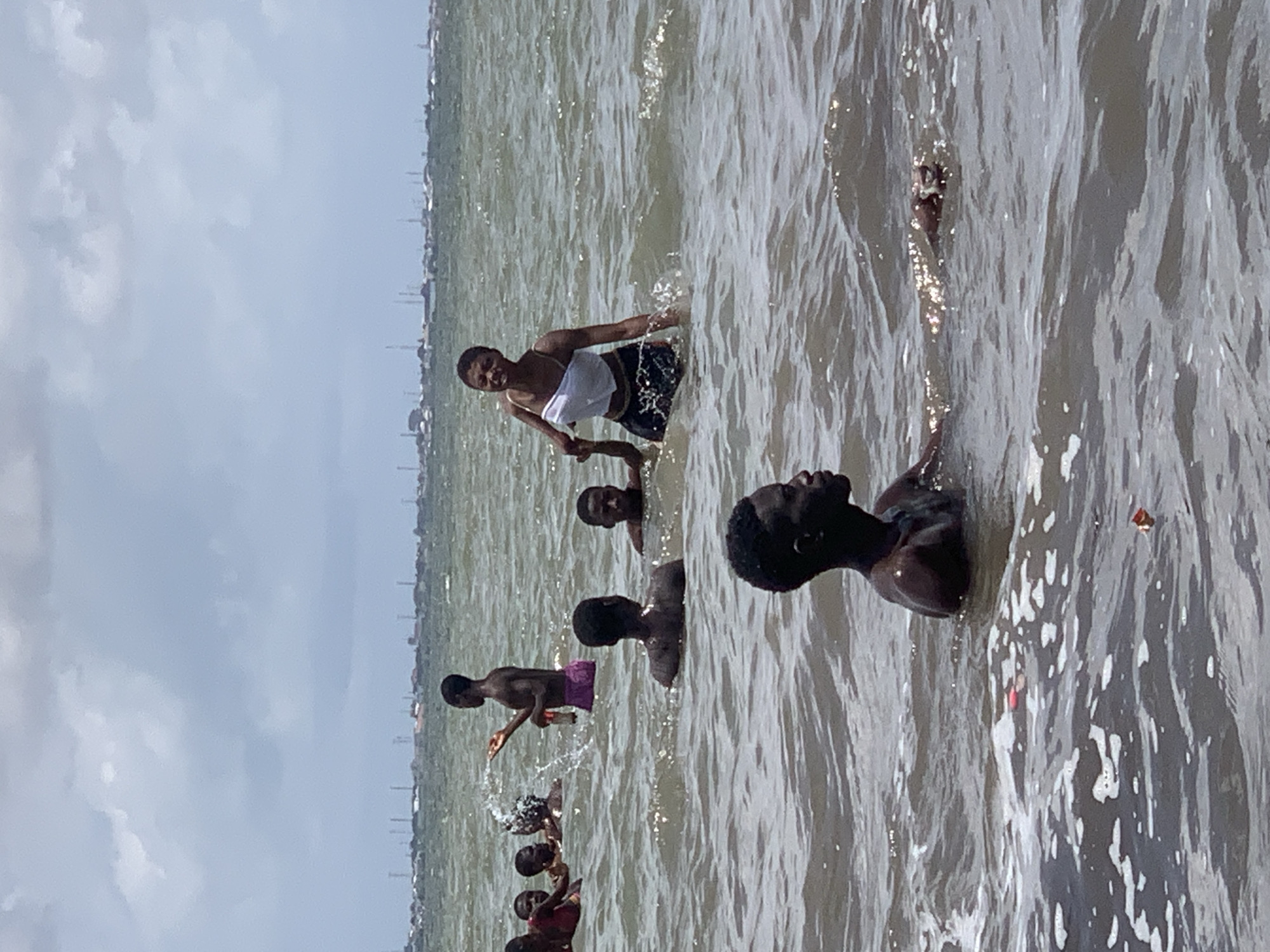 A medium shot of a group of people bathing in water, with a cityscape visible in the background. The water appears murky. From foreground to background: A group of people are partially submerged, with their heads and shoulders visible; one person is facing to the right with their head above water; one person is throwing water towards the left. Another person is standing and facing to the left with a white cloth on their torso.
It appears to be a beach in Ikorodu, Nigeria.