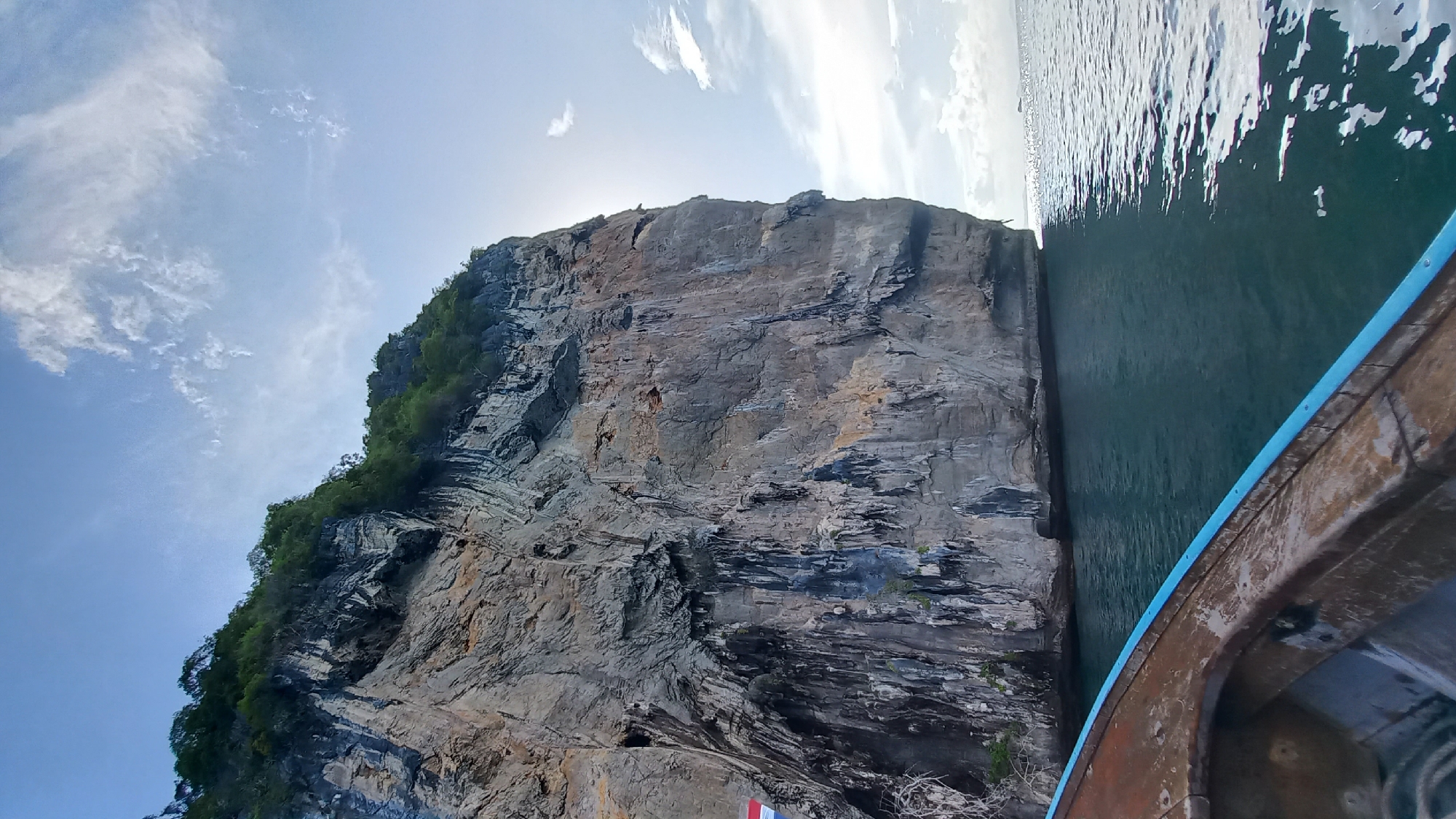 The image shows a view from a boat next to a tall rock cliff in Ban Ao Nang, Thailand. The cliff is predominantly gray with areas of vegetation. The sky is pale blue with scattered clouds. The sea is calm with shimmering sunlight. The boat's side, a combination of brown wood with blue trim, is visible.