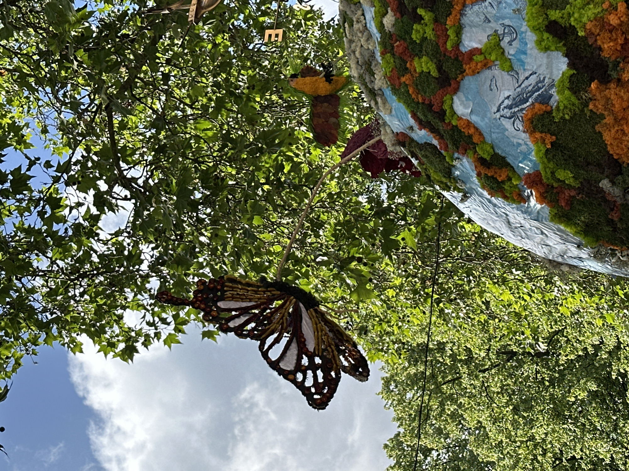 The image is a low-angle shot looking up into the foliage of trees in London, United Kingdom. The sky is visible in the lower left, showing some blue sky and white clouds. Hanging from the trees are decorative objects, including a large, stylized butterfly with brown and white patterned wings. Also visible is a moss-covered globe-like structure with shades of green, orange, and a white and blue base with an anchor drawn onto it. The letter "E" hangs in the upper center of the image. The foliage is dense and vibrant green, illuminated by sunlight.