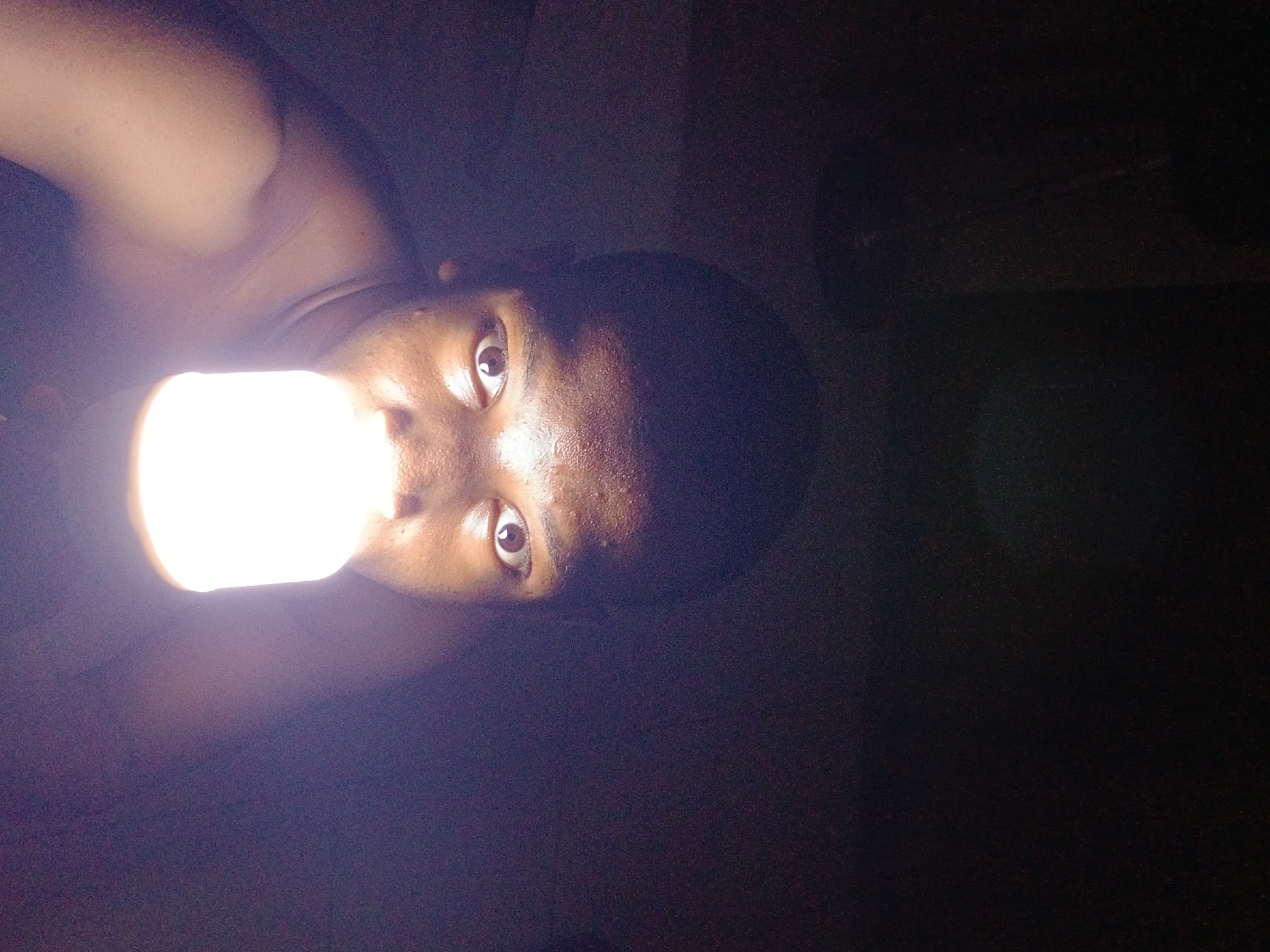 A person is depicted in a close-up, sideways portrait, holding a very bright light source, likely a light bulb or a powerful flashlight, directly in front of their face. The light is intensely white and illuminates the left side of their face, creating strong highlights and deep shadows on the right. The person has dark skin, and their eyes are wide open, looking directly towards the viewer or camera. Their face shows a neutral or focused expression, with some shine evident on their forehead and cheek, possibly due to sweat or the direct reflection of the light. Only the person's head, part of their shoulder, and an arm holding the light are visible. The background is almost entirely black, indicating a very dark environment, such as a room at night or a deeply shadowed area. The extreme contrast between the bright light and the surrounding darkness is a prominent feature of the image. No discernible activity beyond posing with the light source is evident. There is no visible text in the image. The scene is set in what is externally identified as Lagos, Nigeria.