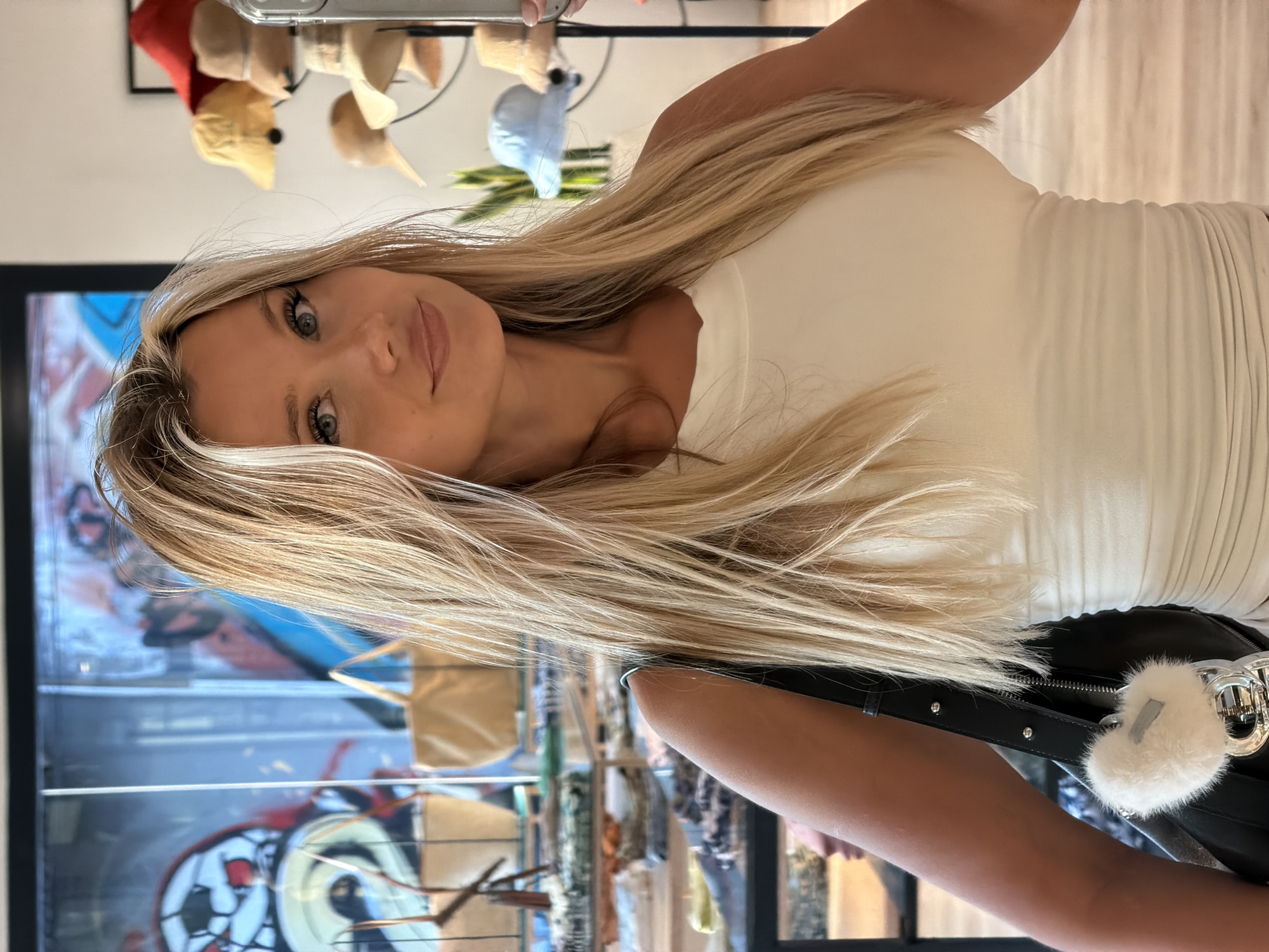 A young woman with blonde hair and blue eyes is standing in front of a display of hats and handbags. She is wearing a white tank top and a black purse with a white fuzzy keychain. The background shows a store interior with shelves of merchandise and colorful graffiti on a wall. She is holding a phone in her right hand and taking a selfie in a mirror.