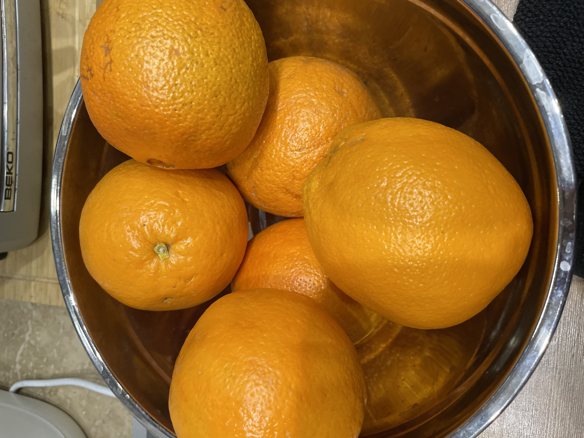 A top-down shot shows a metal bowl filled with six oranges. The bowl sits on a light-colored surface with a visible kitchen appliance to the left and a dark-colored object, possibly fabric, to the right. The oranges are of various sizes and show slight surface imperfections. The interior of the bowl reflects light, indicating a glossy surface. The location in the city of Лесной Пейзаж Кп (шарапово), Russia cannot be determined by the image.