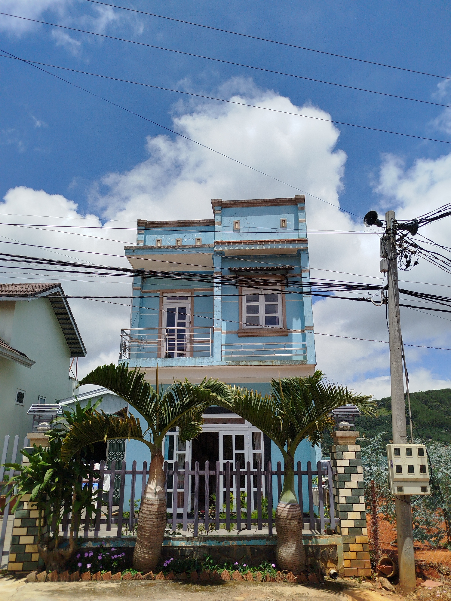 In Trường An, Vietnam, a narrow, light blue, three-story building stands prominently under a bright blue sky with scattered white clouds. The building features balconies and windows with white frames. In front of the building, a dark wooden fence encloses a small yard with two palm trees and some flowering plants. To the right of the building, a concrete utility pole with numerous black wires is visible, along with a white electrical box. Another building, painted in a pale green color, is partially visible to the left of the blue structure. The ground in front of the fence is paved, with a patch of red dirt visible to the right. The scene appears to be a residential area with a view of rolling hills in the background.