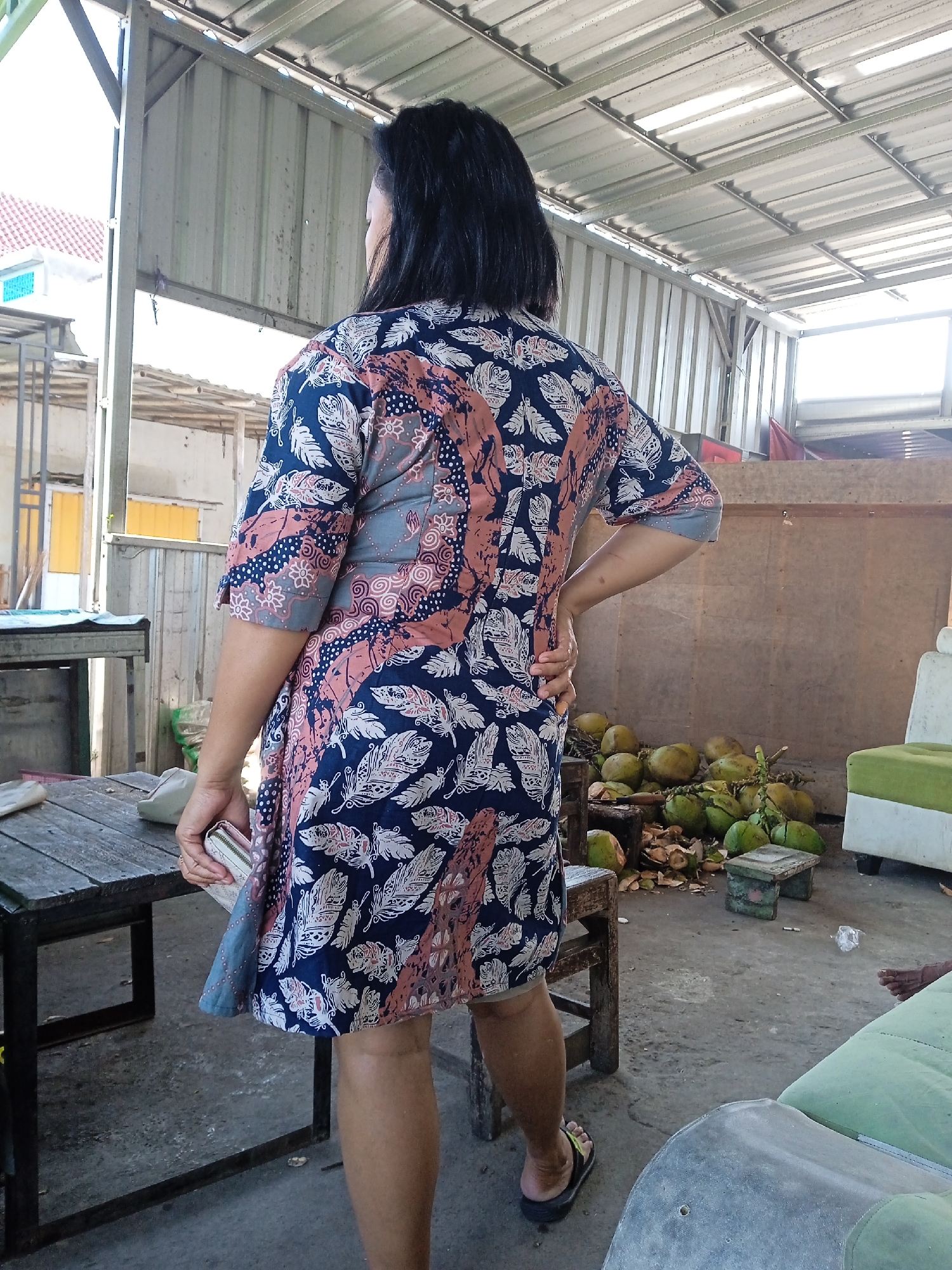 A woman stands with her back to the viewer in what appears to be an open-air structure. She wears a short, patterned dress, and slides on her feet. Her hair is dark. She is holding a wallet or small clutch in one hand and has the other hand on her hip. The structure has a corrugated metal roof supported by metal beams. A wooden table sits to her left, and in the background, a pile of coconuts lies on the concrete floor. Other furniture pieces are also visible, including a light green couch. This scene is in Turunsih, Indonesia.