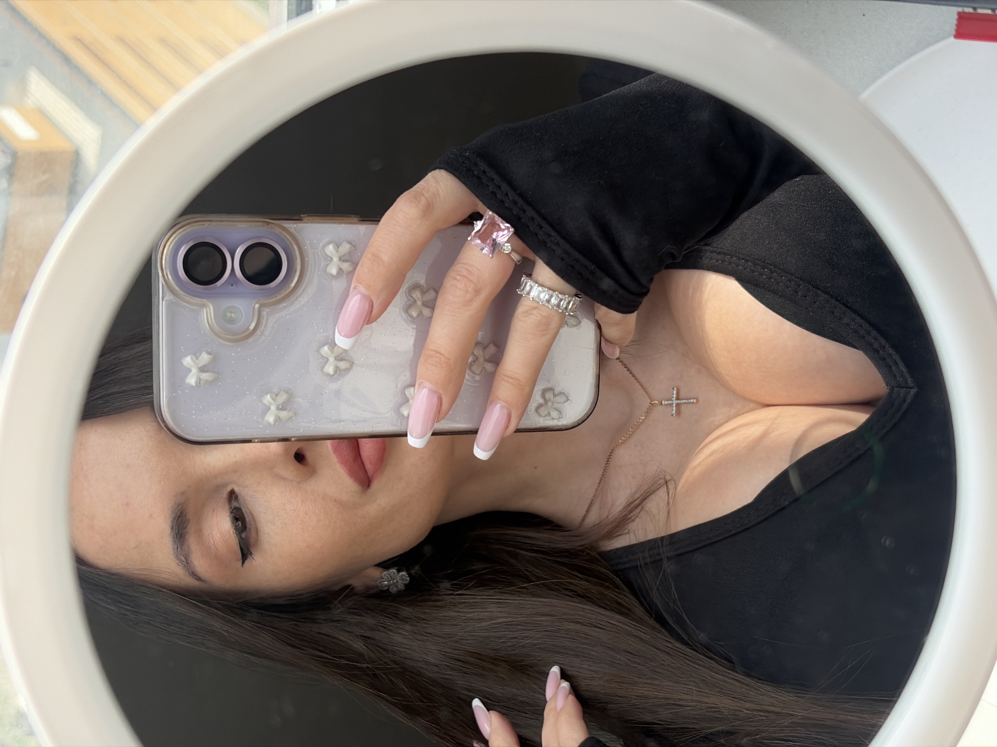 A woman is taking a selfie in a circular mirror, holding a phone in front of her face. She is wearing a black top, a necklace with a cross pendant, and several rings on her fingers. Her nails are manicured with a French tip design. The phone has a clear case with a floral pattern. The background shows a window and a glimpse of buildings. The location is potentially Saint Petersburg, Russia, inferred from the urban setting visible through the window.