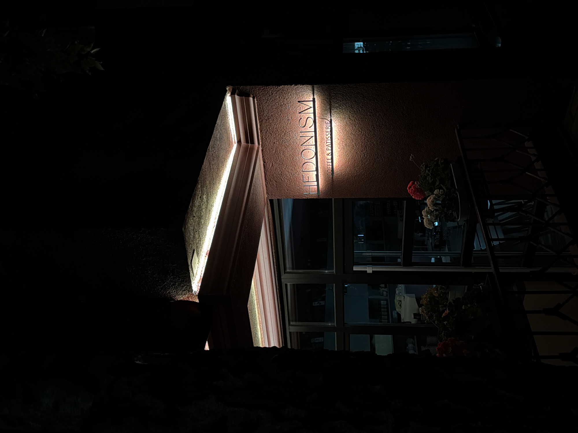At night, a structure illuminated by light is seen. The building displays the word "HEDONISM" with "COFFEE & PATISSERIE" underneath. The location is Unknown, Unknown. A window with plants is visible, along with a metal structure nearby. The background is dark.