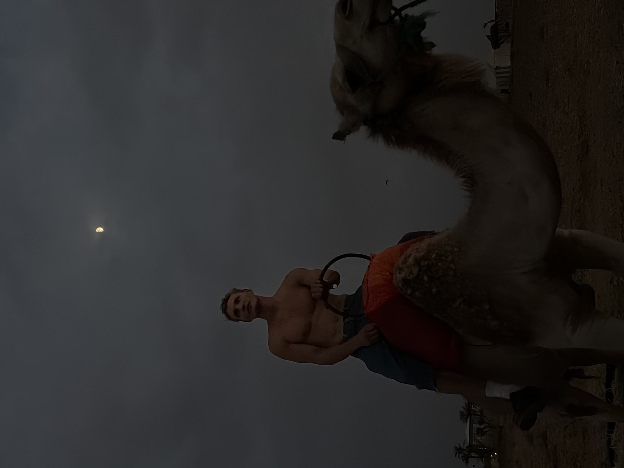 A man with a muscular physique, shirtless and wearing dark shorts and white socks, is seated on a camel. The camel is adorned with a red saddle pad. The man holds onto a dark ring-shaped rein. The scene takes place in Douar Ait Daoud, Morocco, under a dark, cloudy sky with a sliver of moon visible. The ground appears to be sandy or dusty. In the background, there are tents and silhouettes of palm trees, suggesting a desert or oasis setting.