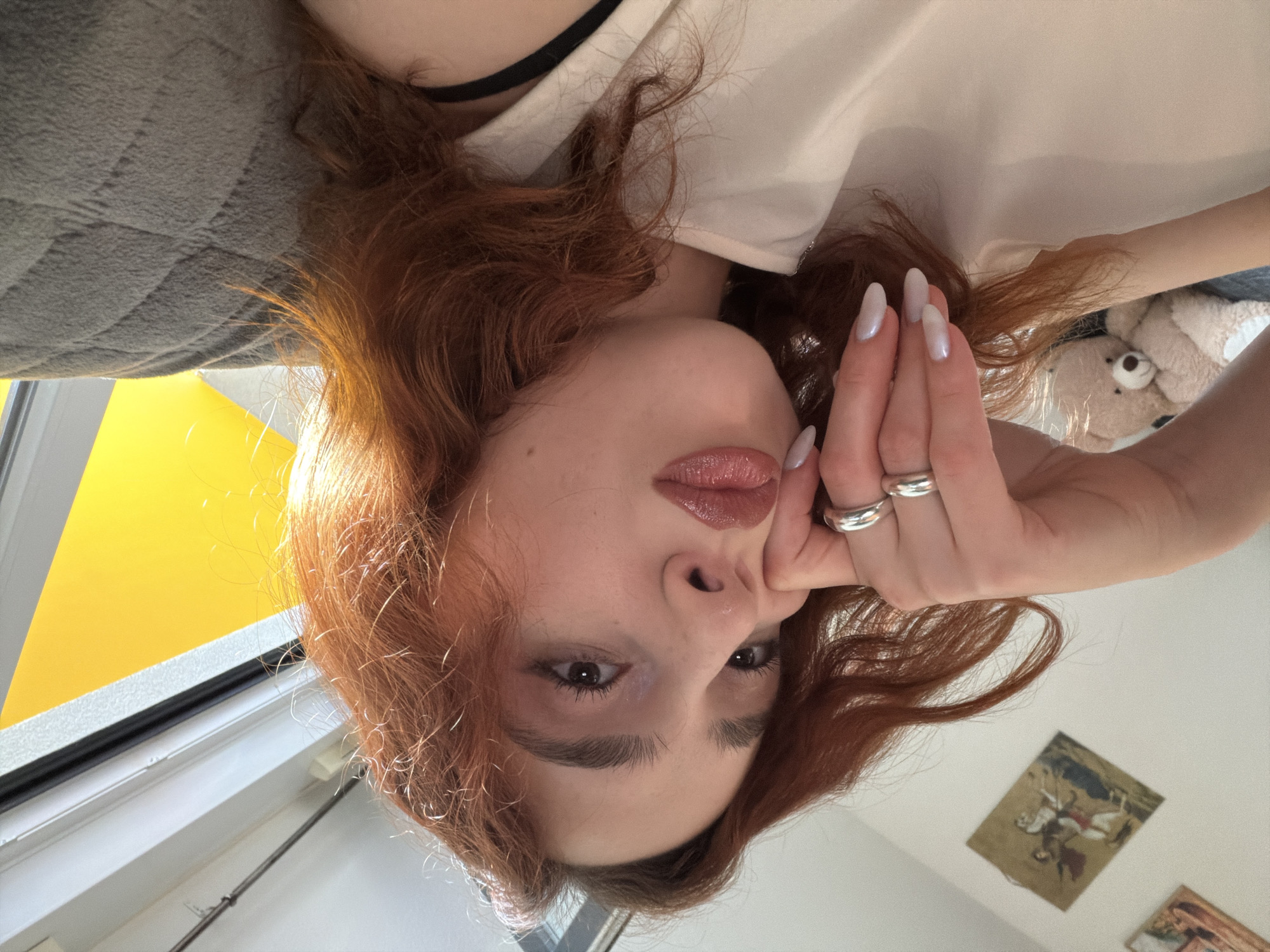 A photograph displays a person with red hair, fair skin, and neutral expression. They are adorned with rings on their left hand, which is positioned near their face. The background includes a gray, quilted surface, possibly a blanket or cushion, and a yellow wall segment. The individual is partially dressed in white, with a hint of black attire visible. Additionally, a teddy bear and framed artworks are present in the scene. The location cannot be determined as Strasbourg, France without additional information.