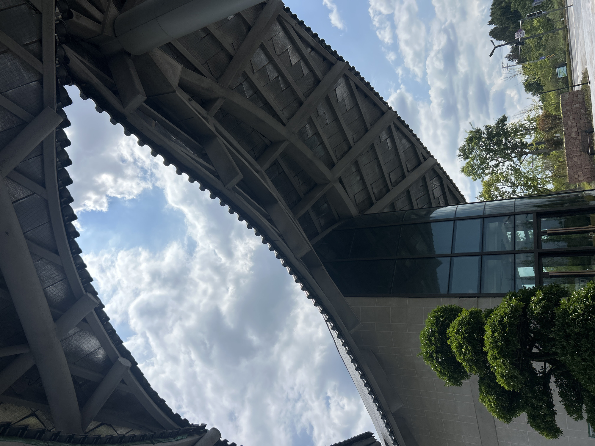 The image shows a view from beneath a large, curved, tiled roof structure with exposed wooden beams. The roof extends over a modern building with a glass facade. To the left of the building, a large, well-maintained, green shrub is visible. The sky above is partly cloudy with a bright blue sky visible through gaps in the clouds. In the background, there are trees and some distant foliage, as well as a traffic light and what appears to be a street. The scene is likely an outdoor public space in Jianyang City, China, possibly a park, plaza, or the entrance to a building. There are no people visible in the image.