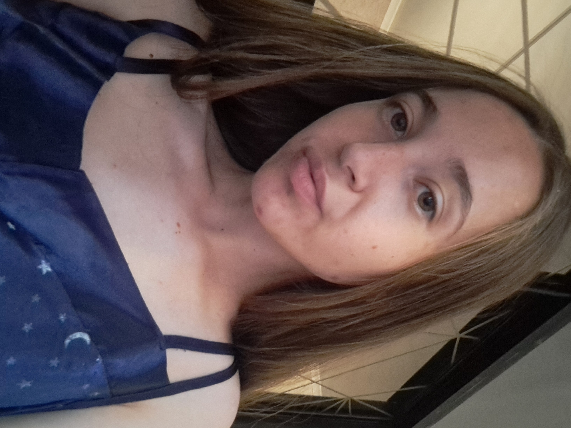 A young woman with shoulder-length brown hair and light skin is shown in a close-up selfie. She is wearing a dark blue spaghetti strap dress with a celestial pattern of stars and a moon. Her head is tilted slightly to her right. The background consists of a light-colored ceiling with visible beams and a dark, lattice-like structure to her right. The image was captured in Vypasne, Ukraine.
