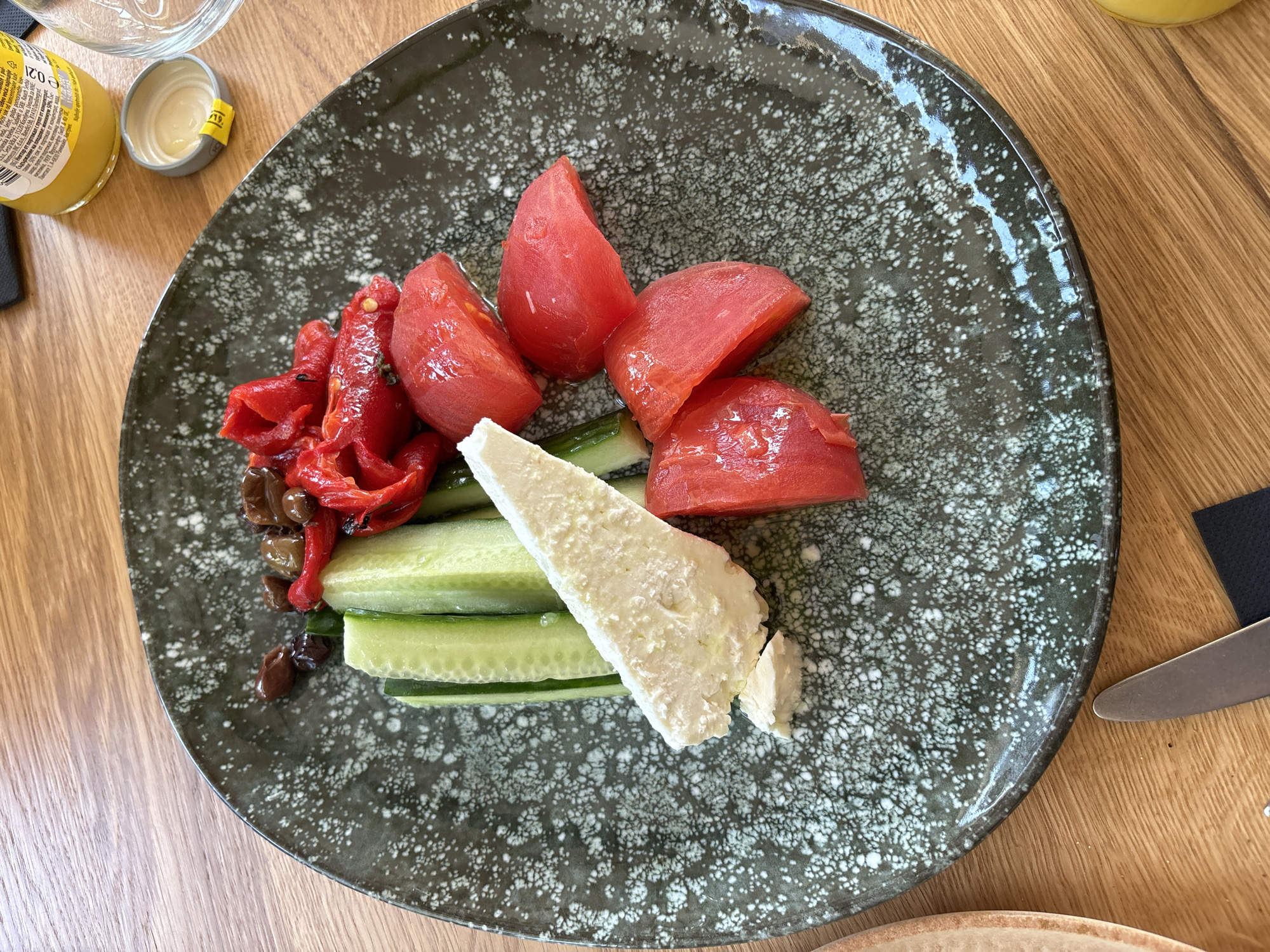 A dark grey-green ceramic plate, featuring a distinct white speckled pattern, is centrally positioned on a light brown wooden table. The plate contains a fresh meal composed of various ingredients. Arranged vertically on the left side are several thick, bright green strips of cucumber. To their right, a substantial wedge of white, crumbly cheese, likely feta, rests partially broken. Further to the right, a cluster of vibrant red tomato wedges, appearing peeled, are visible. Above the tomatoes and slightly to the left, a portion of reddish-orange roasted bell pepper strips is mixed with dark and green olives.
In the upper right corner of the image, a yellow glass bottle with a white label is partially visible. The label features Cyrillic text, clearly stating "Произведено в България" (Produced in Bulgaria). Adjacent to the bottle, a metallic grey bottle cap with a yellow band and some liquid residue sits on the table. A clear drinking glass is partially visible on the far right. In the bottom left, the silver handle of a knife rests on a small black napkin or coaster. The wooden table, with its visible grain, serves as the background for the entire scene. The setting indicates a dining environment, consistent with a restaurant or cafe in Sofia, Bulgaria, given the visible product information confirming Bulgarian origin.