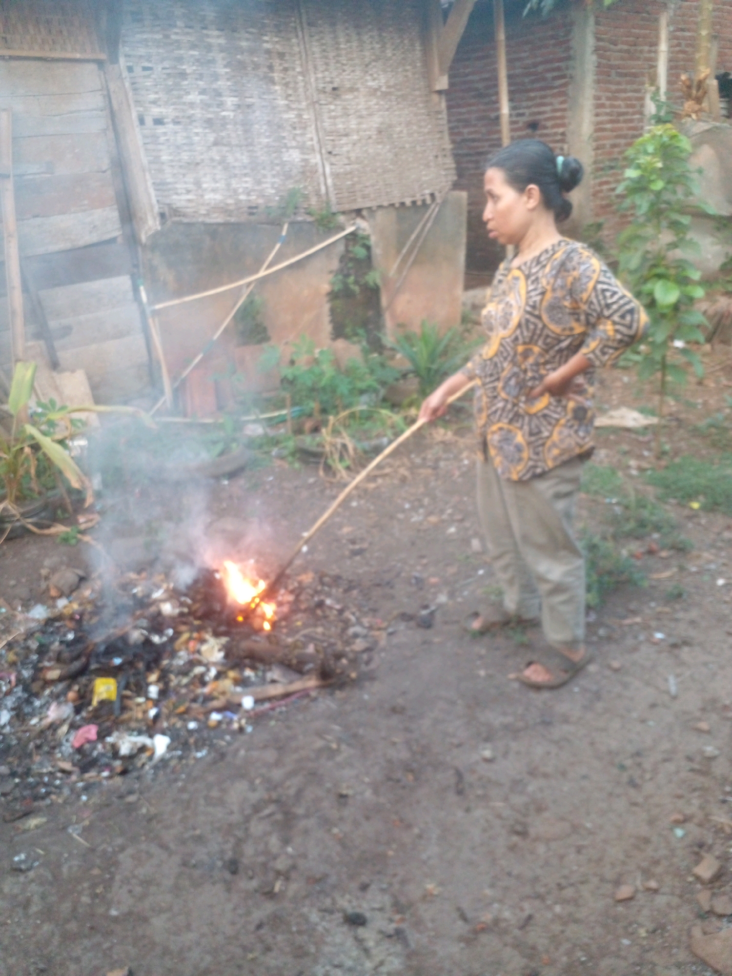 A woman, appearing to be of Indonesian ethnicity with dark hair pulled back in a bun, is standing in what appears to be a rural or semi-rural outdoor setting in Pungangan, Indonesia. She is wearing a patterned long-sleeved shirt with yellow and black designs and light-colored trousers. Her left hand is on her hip, and she holds a long, thin stick in her right hand, which she is using to stir a small pile of burning trash and debris. The fire is producing smoke. The ground is dirt, and there is evidence of refuse and various objects on the ground around the fire. In the background, there are structures that appear to be made of wood and possibly bamboo or woven materials, as well as a brick wall. Vegetation, including green plants and trees, is also visible. The overall impression is that of a domestic or informal waste disposal activity in a modest environment.