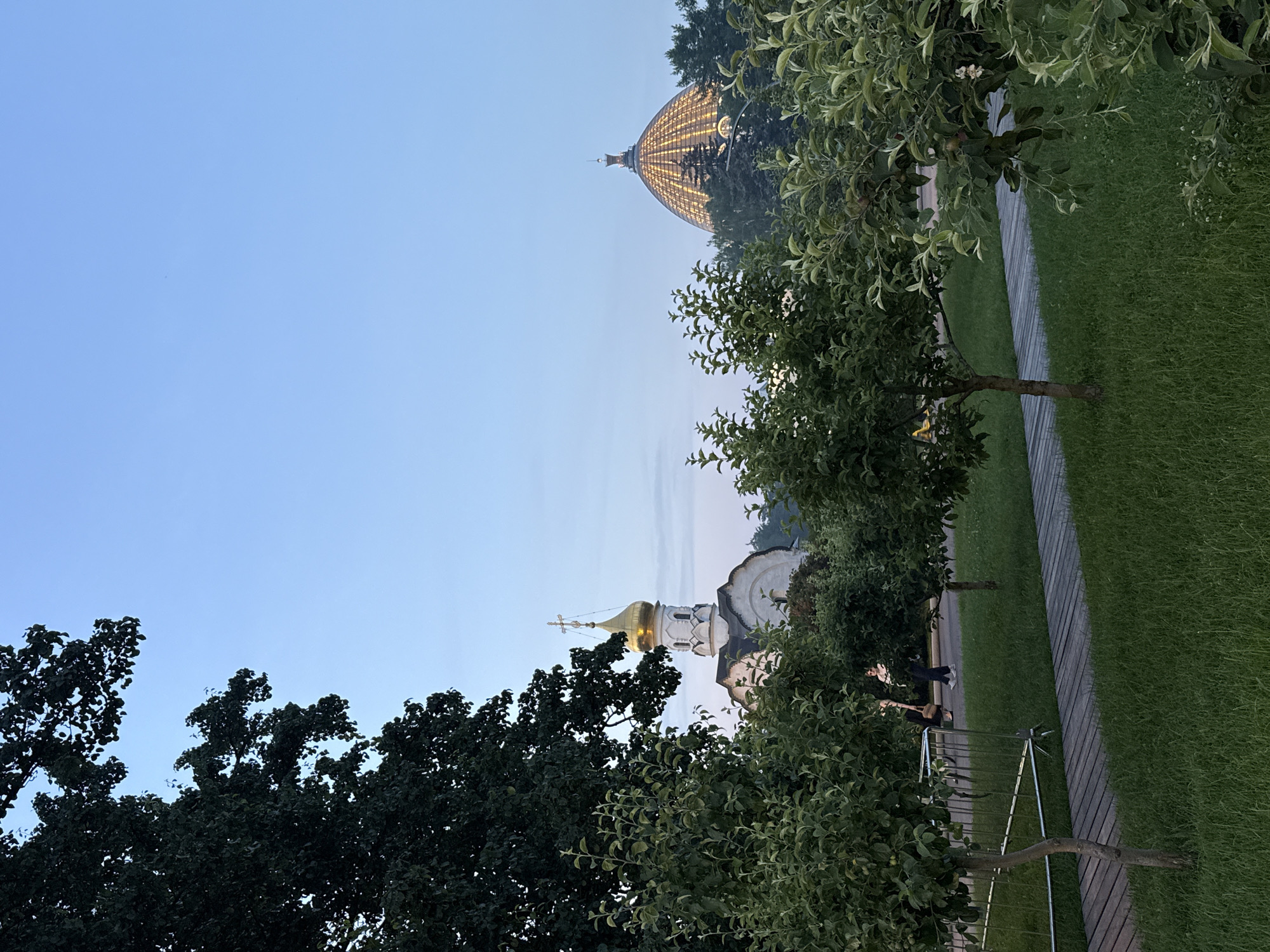 The image depicts a park scene in Moscow, Russia, during dusk. In the background, a golden-domed orthodox church with a cross on top is visible, partially obscured by trees. To the right, a large, illuminated glass dome structure stands prominently, also surrounded by trees. The foreground features a wooden pathway cutting through a lush green lawn. Several small trees are planted along the pathway. Two individuals are seen walking on the pathway in the middle ground, and some temporary fencing is present on the left side of the path. The sky is clear and exhibits a gradient of blue, transitioning to a lighter hue near the horizon.