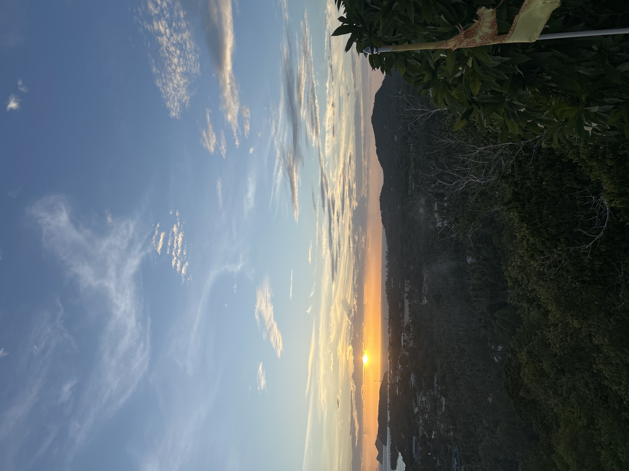 This image captures a sunset over a lush, tropical landscape in Ko Pha-ngan District, Thailand. The sun, a vibrant orb, is low on the horizon, casting a warm, golden glow across the sky and the sea. Wispy clouds streak across the clear blue sky, illuminated by the setting sun. Below, a dense canopy of green vegetation covers rolling hills, suggesting a mountainous terrain. In the distance, a body of water, likely the ocean, meets the land, with a few islands visible on the left. A small settlement with buildings is nestled amongst the trees. In the foreground, the dark silhouettes of trees and branches frame the scene, with a tattered yellow flag on a pole visible on the right. The overall impression is one of natural beauty and tranquility, characteristic of a tropical island at dusk.