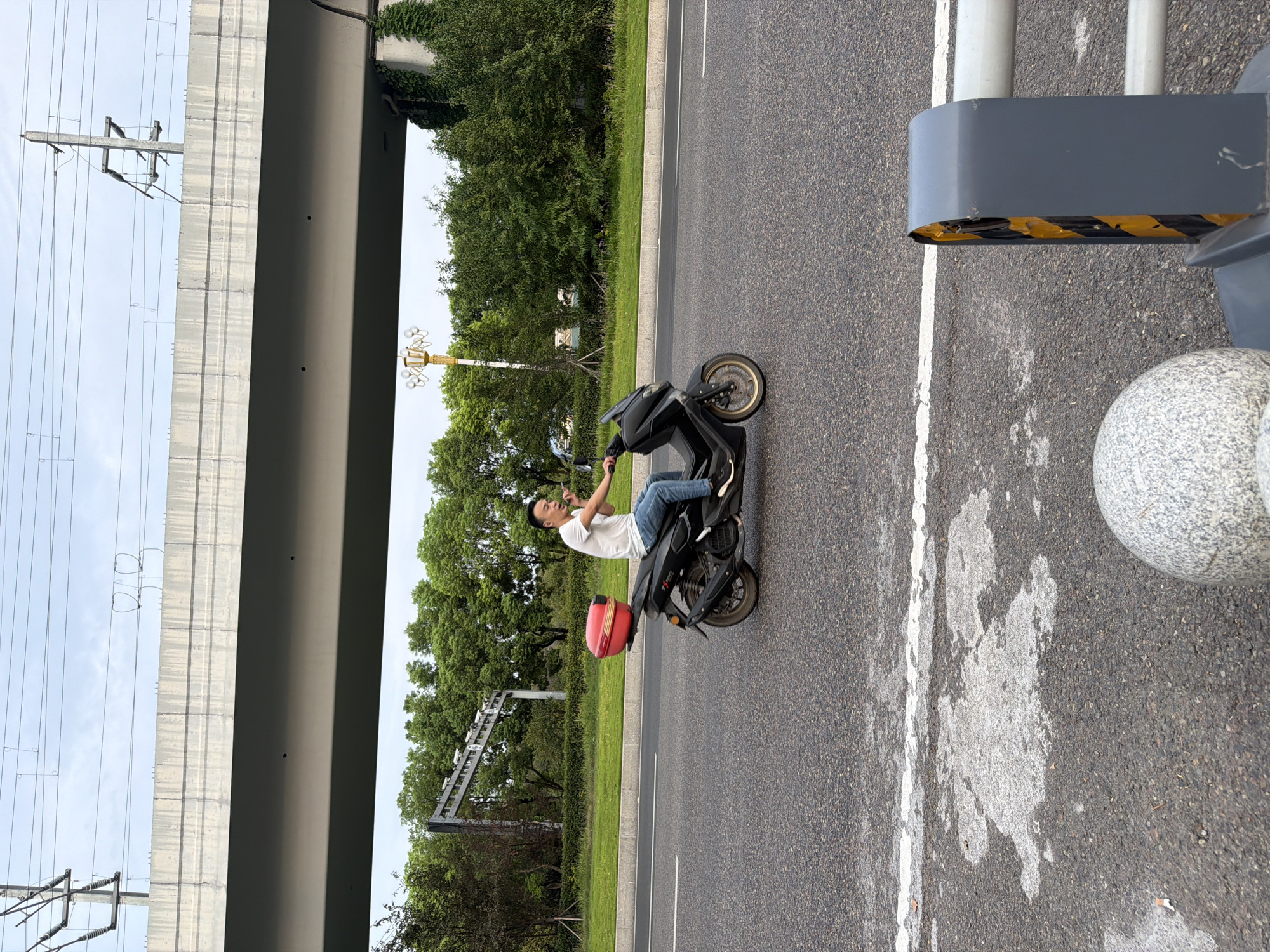 An adult male in a white t-shirt and blue jeans rides a black scooter with a red storage box on the back. He is looking towards the right and speaking with his right hand gesturing. The scooter is on an asphalt road with a white line marking the lane. Behind the road is a grassy area with trees and an overpass. Overhead, there are power lines and a cloudy sky. In the foreground, part of a granite ball and a gray and yellow barrier are visible. The image appears to be taken in the city of Jiangjin, China, indicated by the distinct architectural style of the overpass and streetlights often found in the region.