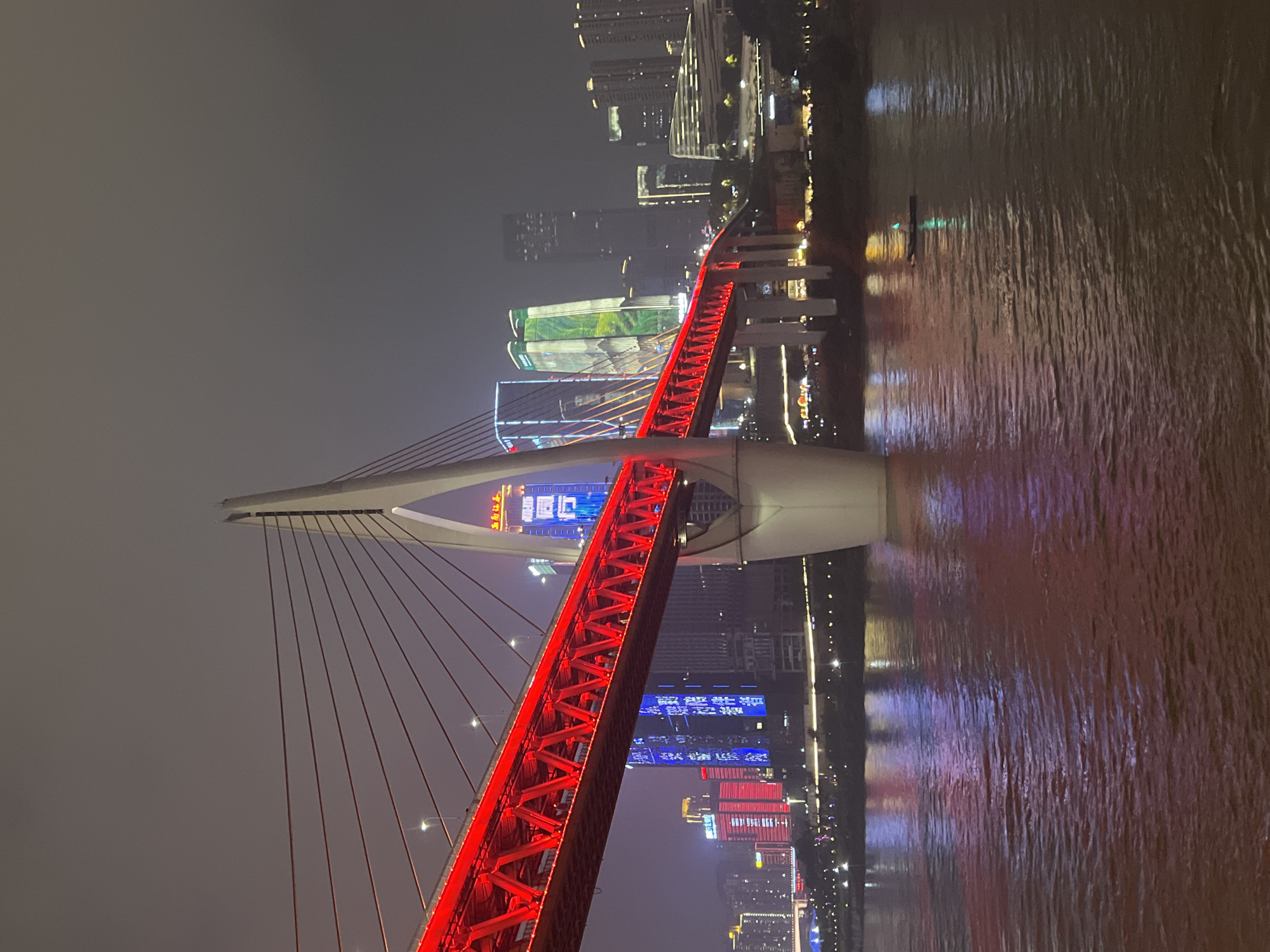 An illuminated cable-stayed bridge spans a body of water at night in Yuzhong District, China. The bridge's deck is a vibrant red, supported by a large white pylon, with numerous red cables extending upwards. The surrounding cityscape is lit with various colorful lights from tall buildings, some displaying blue and green illumination. Reflections of the bridge and city lights shimmer on the dark water's surface. A small dark boat with a green light is visible in the middle of the river. The sky is overcast and dark.