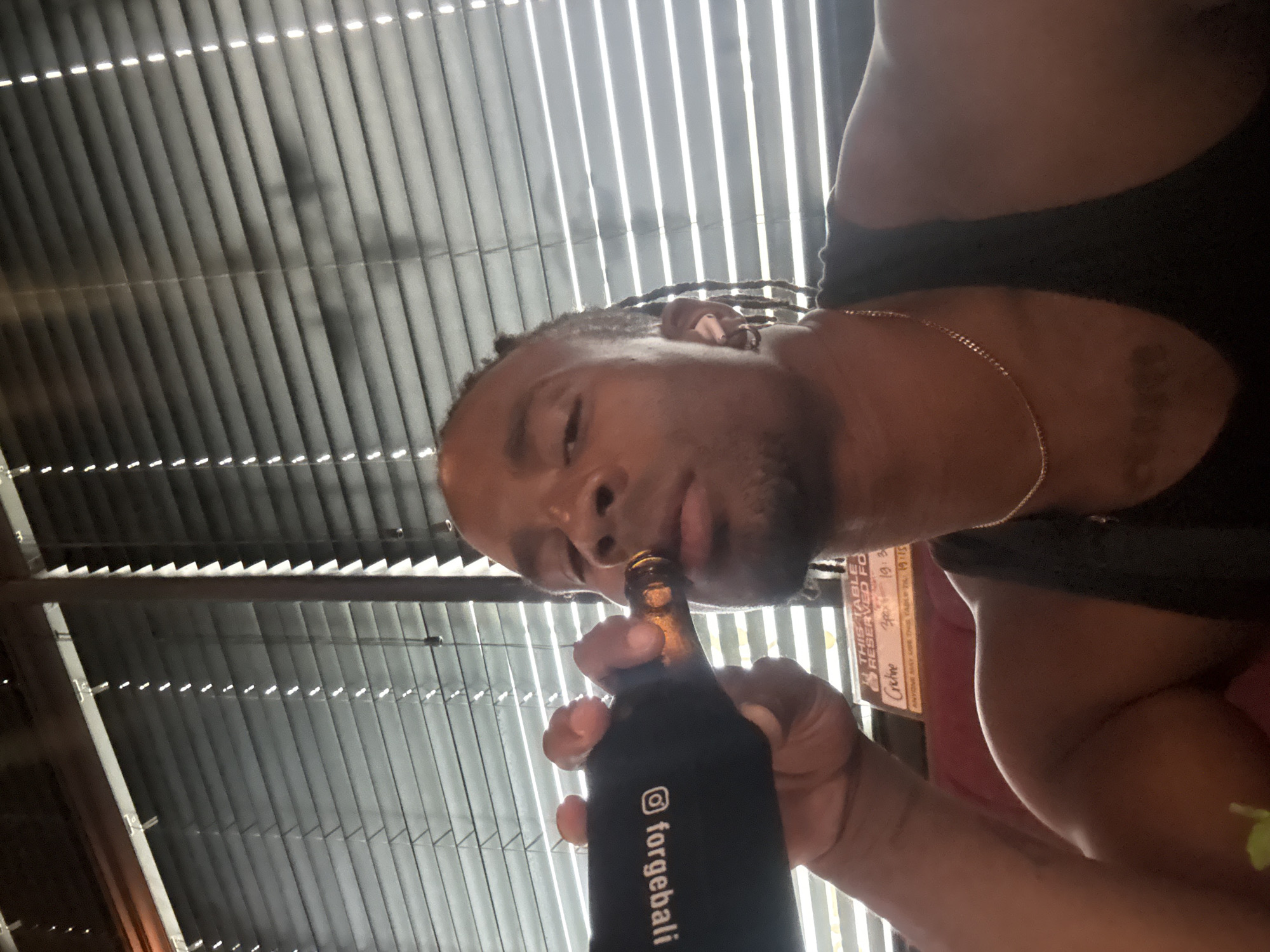 Here is a forensic description of the image.
A man with braided hair and a gold chain is drinking from a beer bottle in a black koozie labeled "@forgebali." He is wearing a black tank top. The background is comprised of window blinds and a sign that reads "TABLE RESERVED FO". Below that is some additional writing that is blurry, but the phrase "ANYONE MAY USE THIS TABLE TIL" is discernible. The sign also states "Cretine," "30. 19.3," and "19.15". The location appears to be a business establishment in the city of Unknown, Unknown.