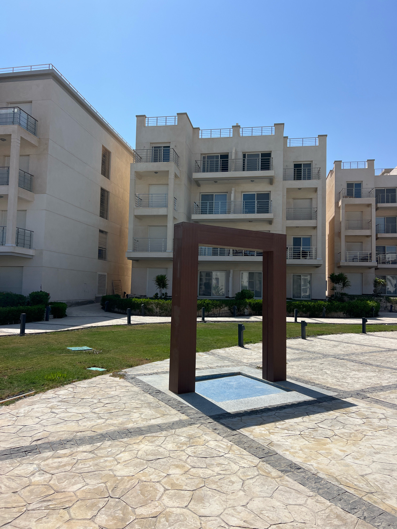 The image captures a clear, sunny day in Sharm El Sheikh, Egypt, showcasing modern residential buildings and an outdoor plaza. In the foreground, a large, dark brown, rectangular wooden frame stands centrally, framing a small, shallow, tiled water feature with a light blue mosaic pattern. The plaza is paved with light-colored, irregular-shaped stone tiles, bordered by a darker band of smaller square tiles. Black bollard-style lights are spaced along the edge of a well-maintained green lawn to the left.
Behind the plaza, two multi-story residential buildings with cream-colored facades dominate the background. The buildings feature multiple balconies with metal railings and large sliding glass doors, many of which are covered by roller blinds. The architecture suggests a contemporary resort or apartment complex. One building on the left is taller and appears to be connected to another section. The building on the right is slightly shorter and is flanked by another similar structure further in the distance. Lush green bushes and small trees are planted along the base of the buildings and around the plaza, adding a touch of nature to the urban environment. The sky is a bright, cloudless blue, typical of the region. No people are visible in the scene.